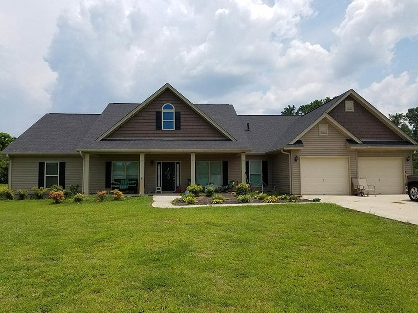 Two-story house with gray siding, white trim, and gabled roof, front porch with lawn chairs, manicured green lawn, concrete walkway, large windows, partly cloudy sky
