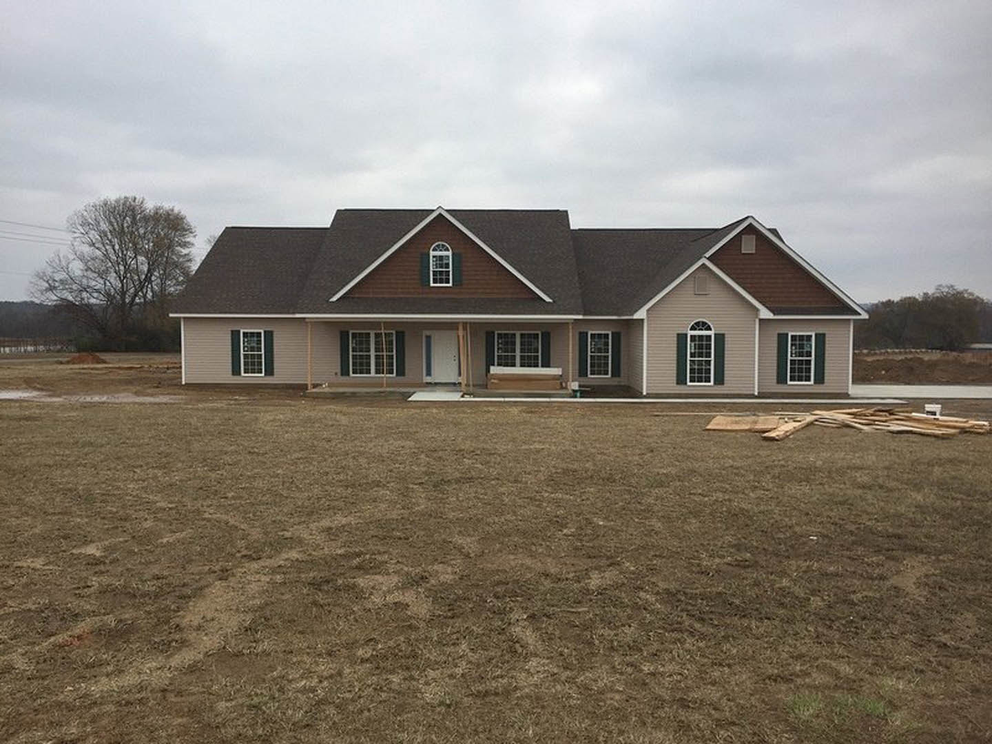 Two-story cottage with white-framed windows, white front door, covered porch, large brown grass yard, leafless tree, and stacked firewood in foreground under cloudy sky