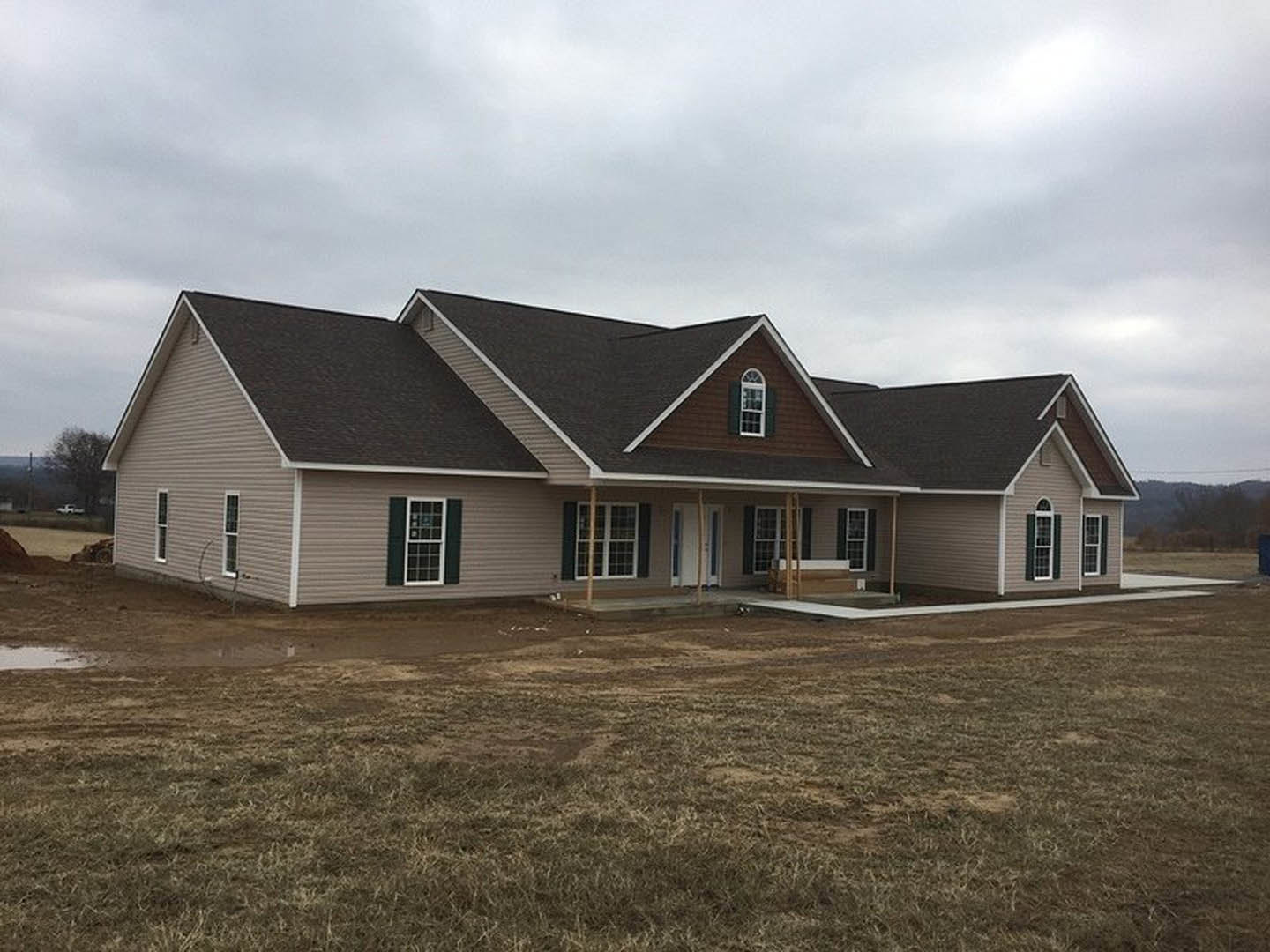 Two-story house under construction with white-framed windows, covered porch, gray siding, and shingle roof, surrounded by dirt field and patchy grass under cloudy sky