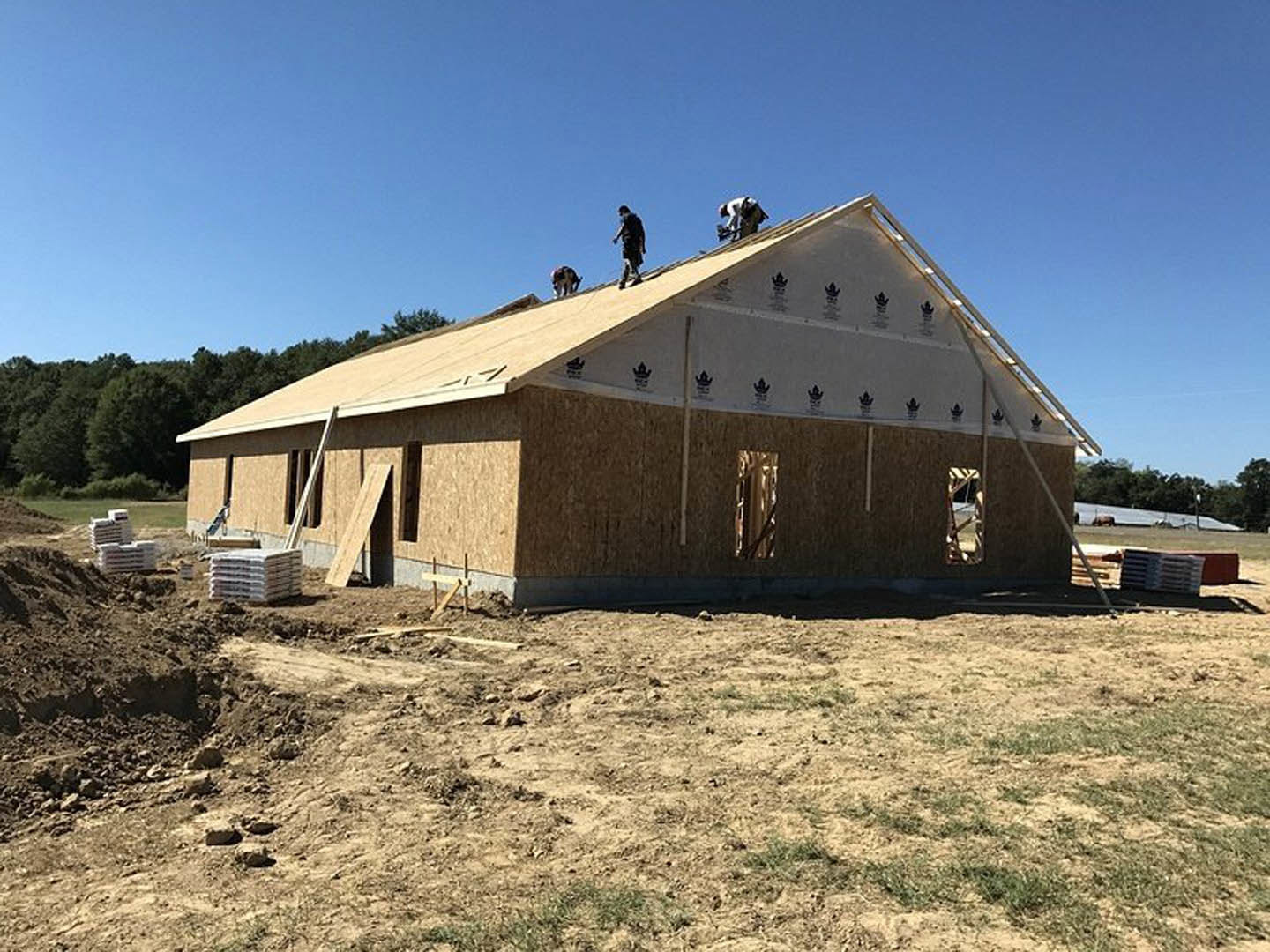 Wood-framed house under construction with workers on the roof, exposed beams, dirt lot in foreground, clear blue sky overhead