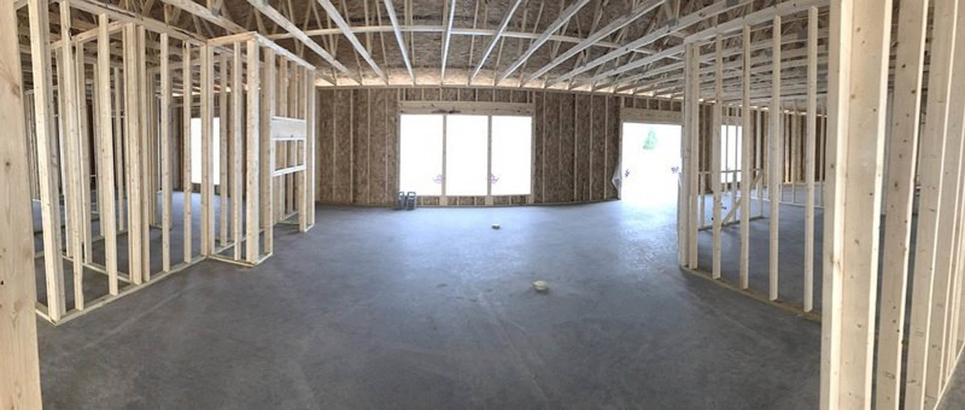 Spacious room with concrete floor, exposed ceiling beams, large windows set in a white wall, natural daylight streaming in, minimal objects scattered on the floor