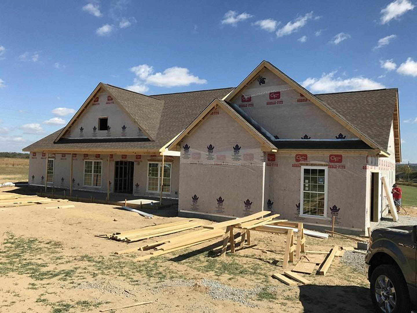Framed house under construction with exposed wood beams, scattered lumber on dirt ground, glass window installed, black truck tire visible nearby