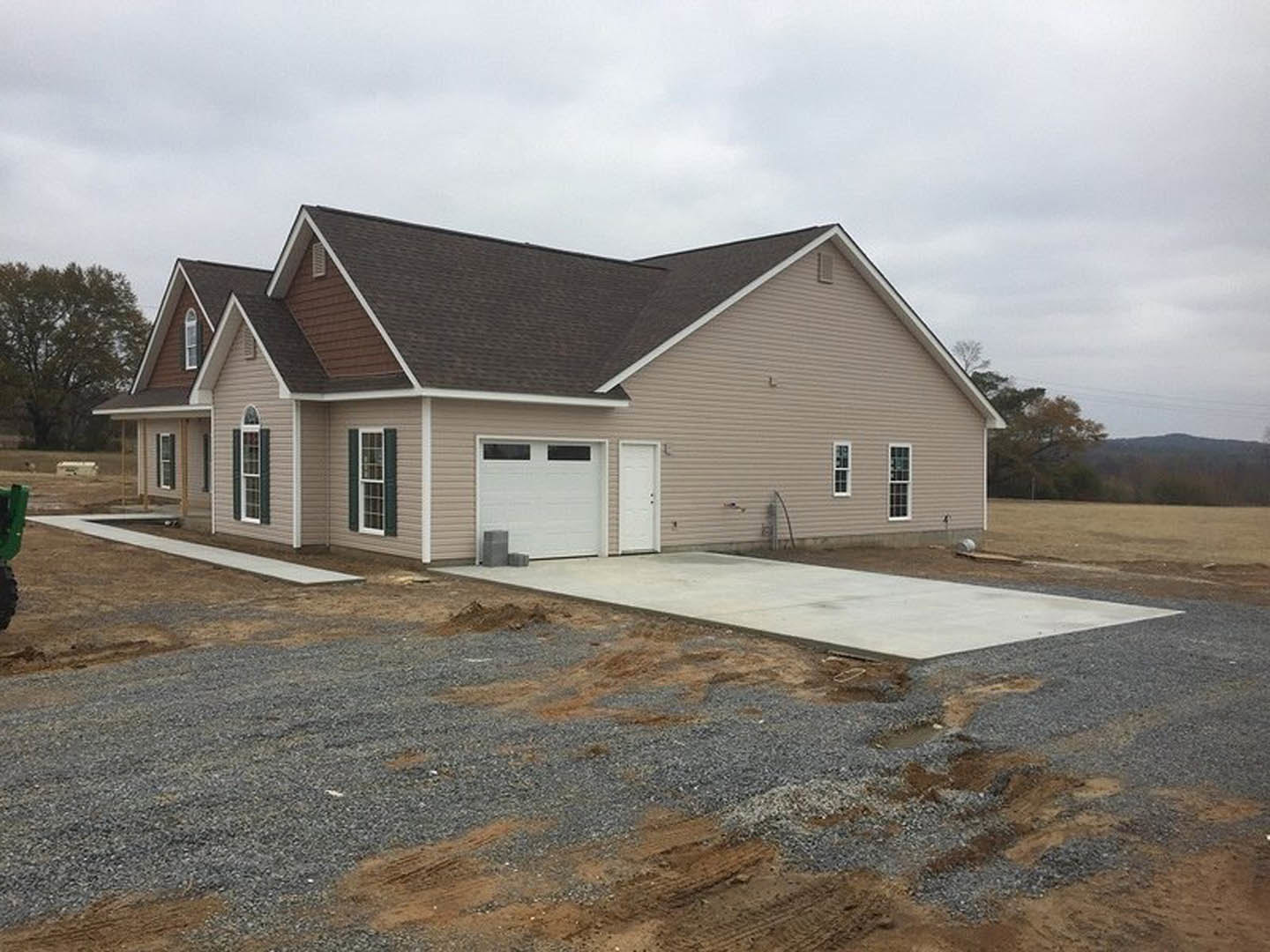 White garage door with upper windows, concrete driveway bordered by gravel, single-story house with light siding, front window, leafy tree in background, partly cloudy sky