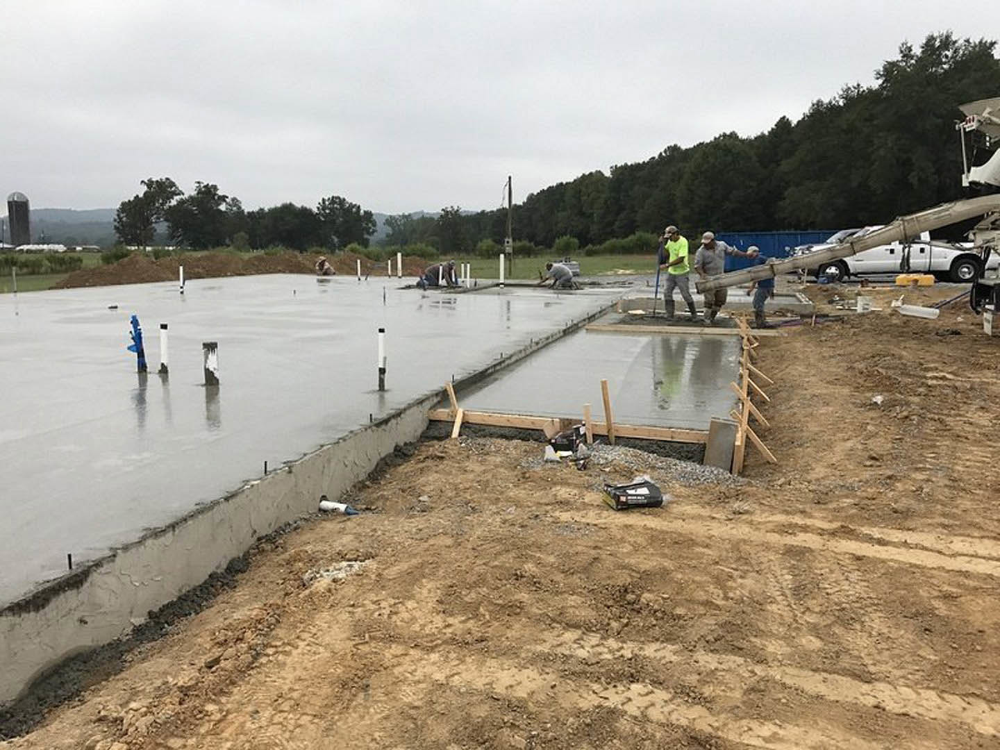 Workers installing concrete slab foundation outdoors, surrounded by construction materials, white truck with black straps, trees, and cloudy sky in background.
