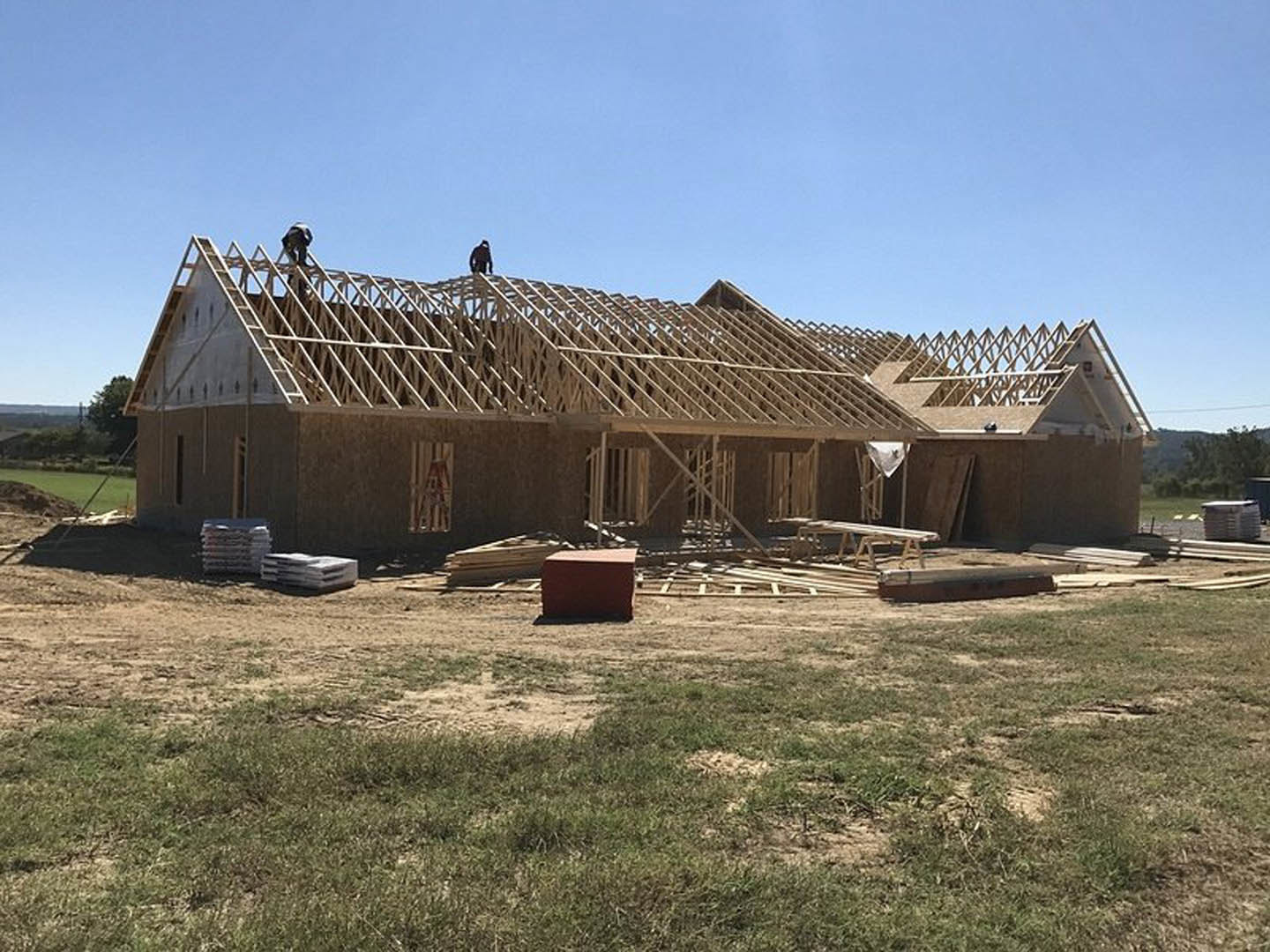 Framed house under construction with workers on roof, surrounded by grassy lot and clear sky