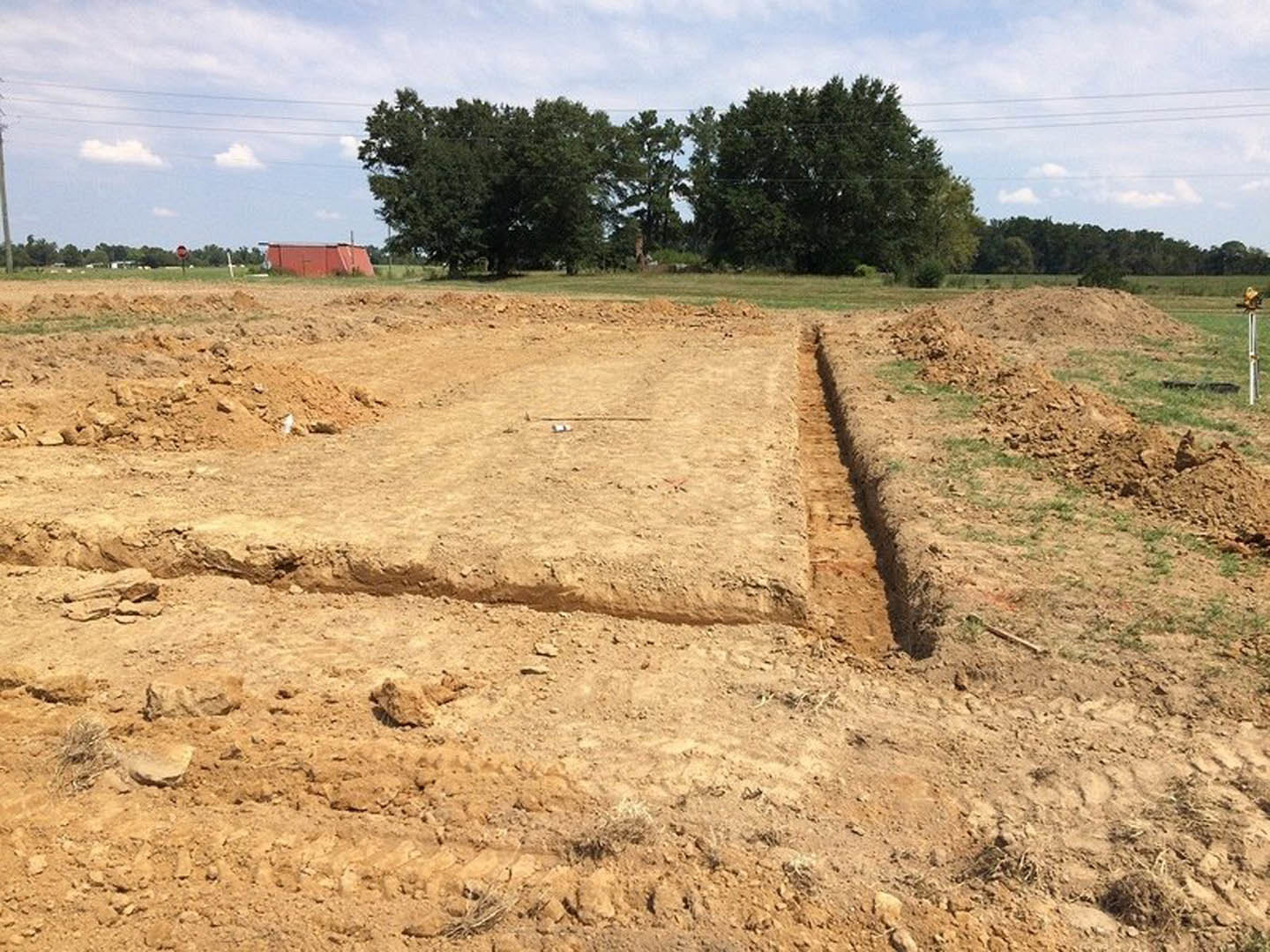 Dirt field with a trench and mound, surrounded by grass and scattered trees under a cloudy sky