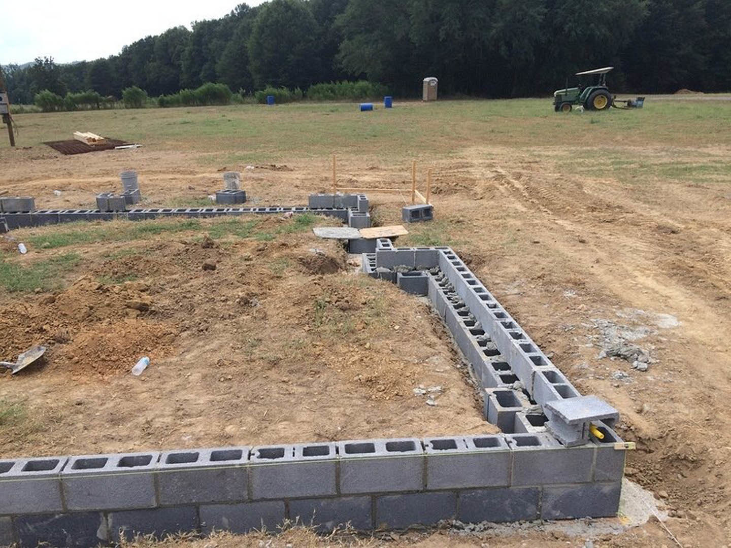 Construction site with exposed soil and foundation blocks, two tractors parked near a partially built brick wall, grassy field and trees in the background under a clear sky