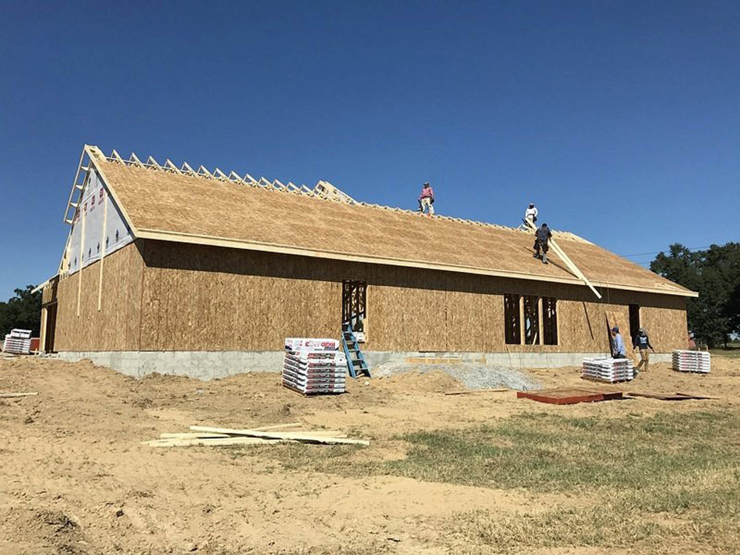 Workers installing roofing materials on a modern cottage-style home with light siding, surrounded by trees and open land under a clear sky