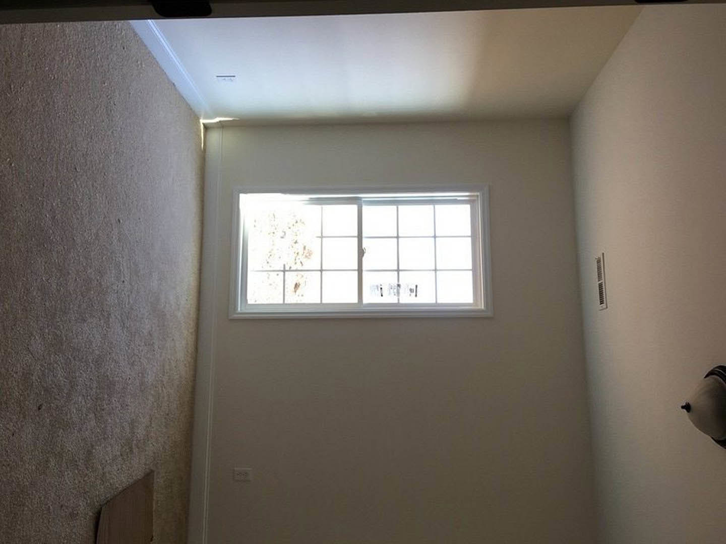 Sunlit room featuring a large window with white blinds, smooth plaster walls, wooden door with metal handle, and ceiling vent.