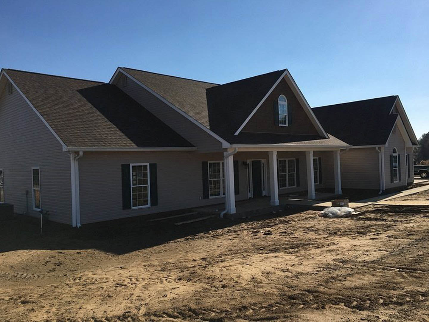 Two-story house under construction with white-framed windows, large covered porch, gray siding, and dirt yard in foreground