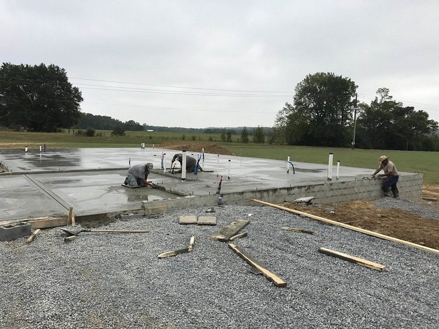 Man kneeling on a newly poured concrete slab near a large leafy tree, with a pile of wood on gravel and scattered lumber in the foreground
