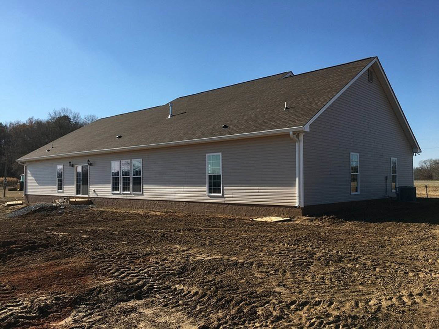Partially built house with shingle roof, multi-pane windows, and light-colored siding, surrounded by bare dirt yard and distant trees under a clear sky