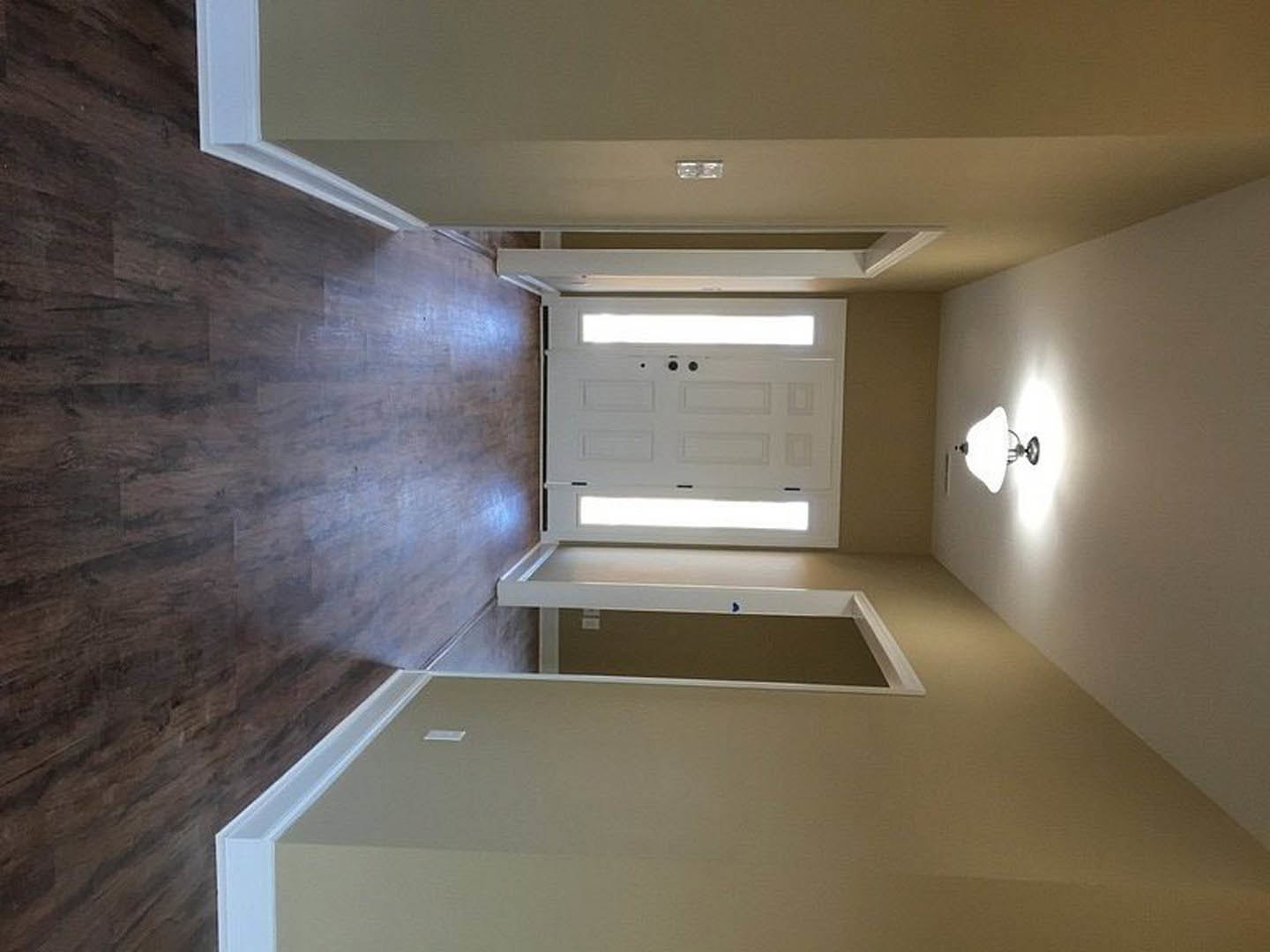 Hallway with white plaster walls, wood flooring, white door, and wall-mounted lamp with white shade