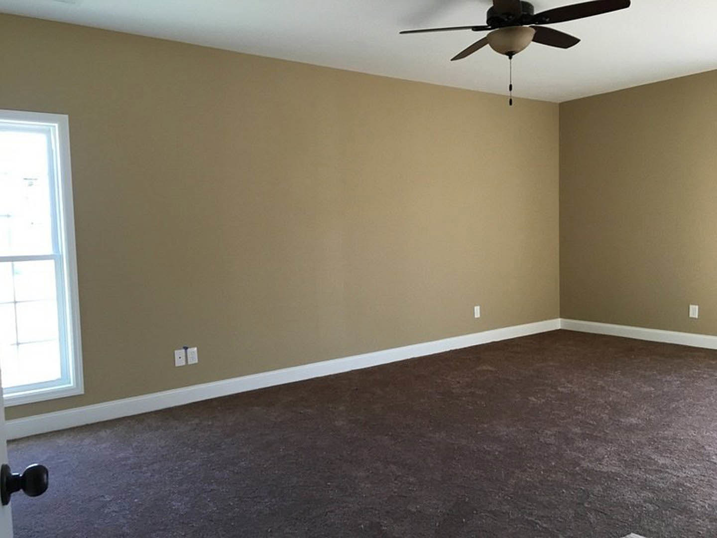 Brown carpeted room with brown painted walls, white electrical outlet, ceiling fan with light fixture, and window with white trim.