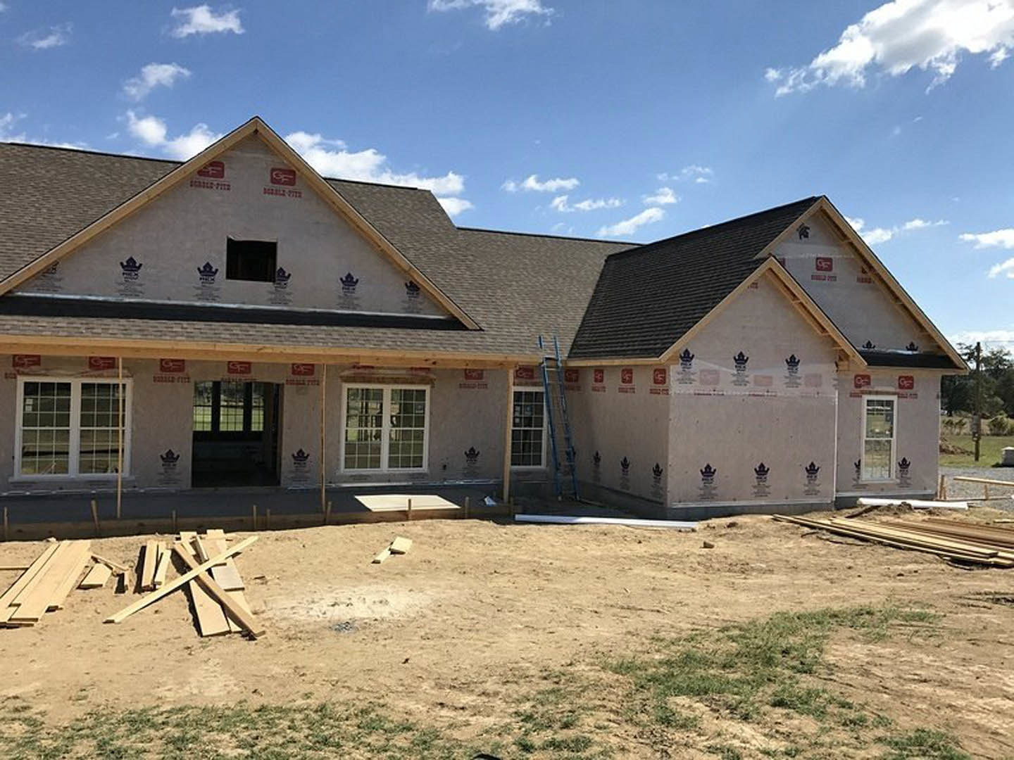 Two-story white house under construction with scaffolding and ladder, multiple square windows with white frames, dirt yard bordered by a fence and patches of grass under a cloudy
