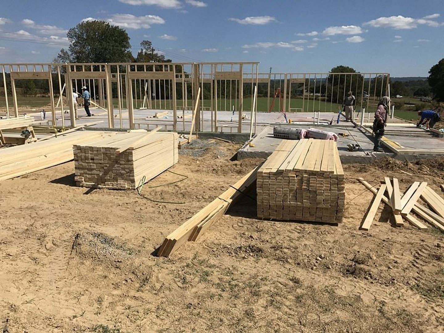 Framed wood beams and stacked planks on a residential construction site, several workers in jackets and helmets, cloudy sky and trees in background