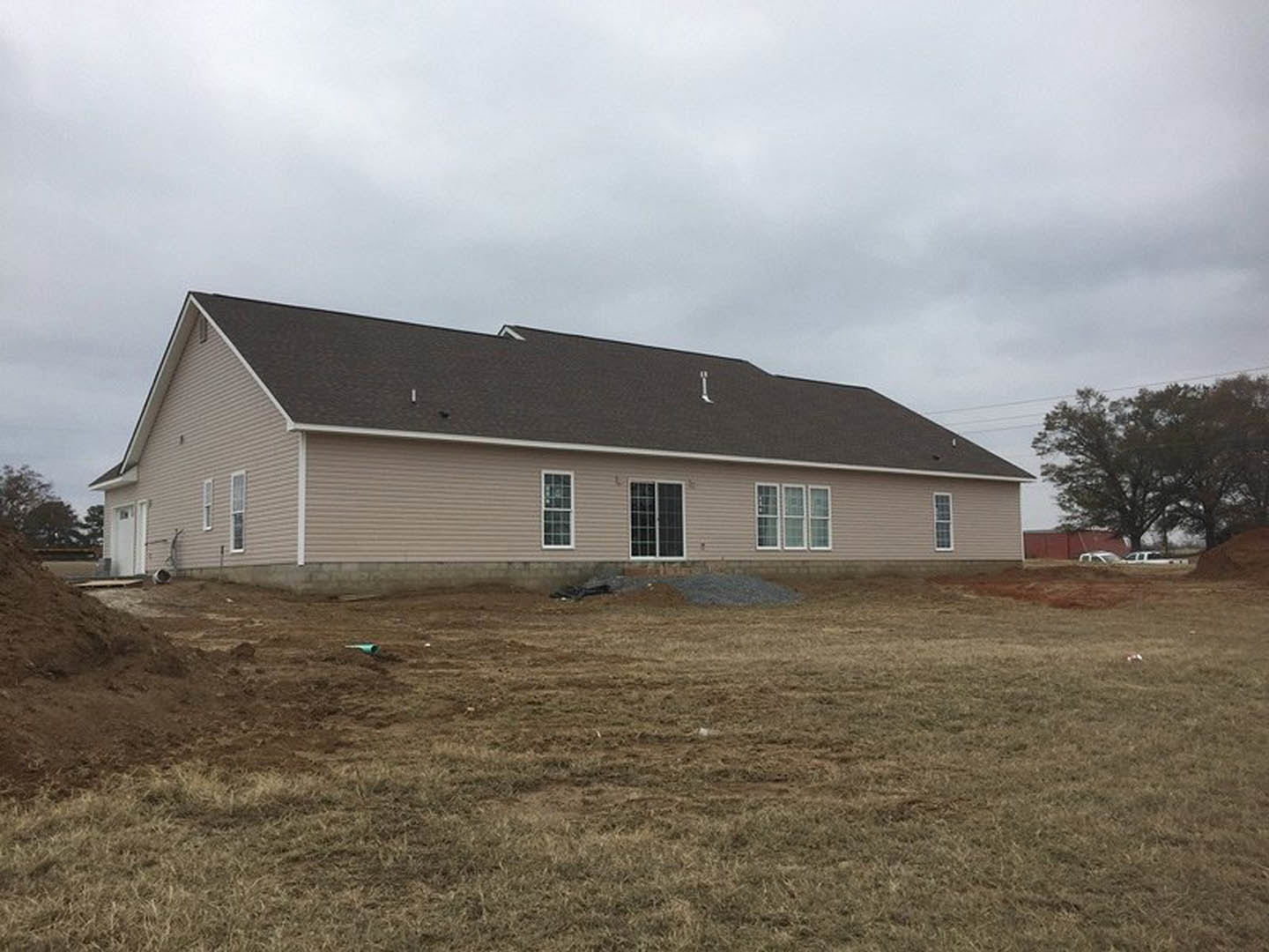 Two-story house with gray roof and white siding, surrounded by a large dirt field, scattered trees, multi-pane windows, and cloudy sky overhead