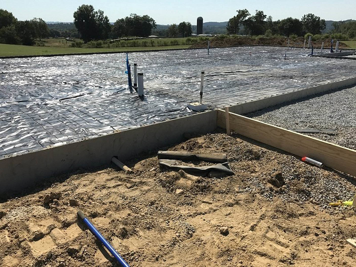 Concrete foundation slab with vertical metal poles, blue pipe emerging from dirt, piles of soil, and surrounding grassy field with trees in background