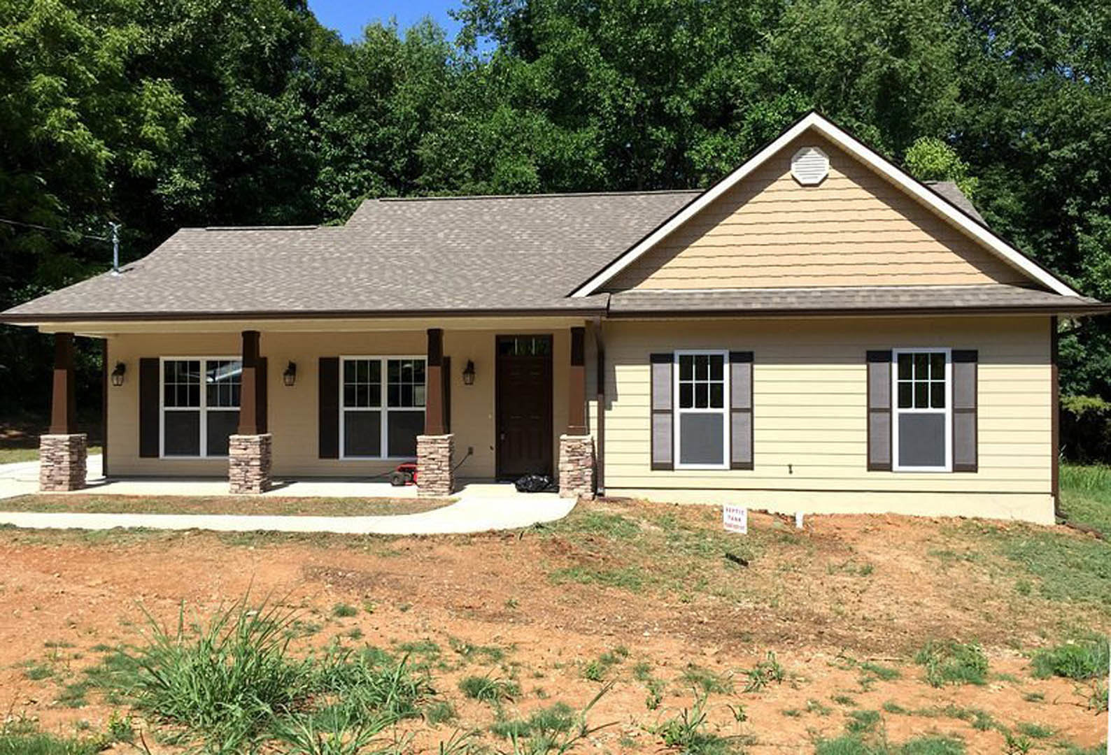 Modern house exterior with black front door bordered in white, large windows, covered porch, and dirt yard with sparse plants and a lawn mower.