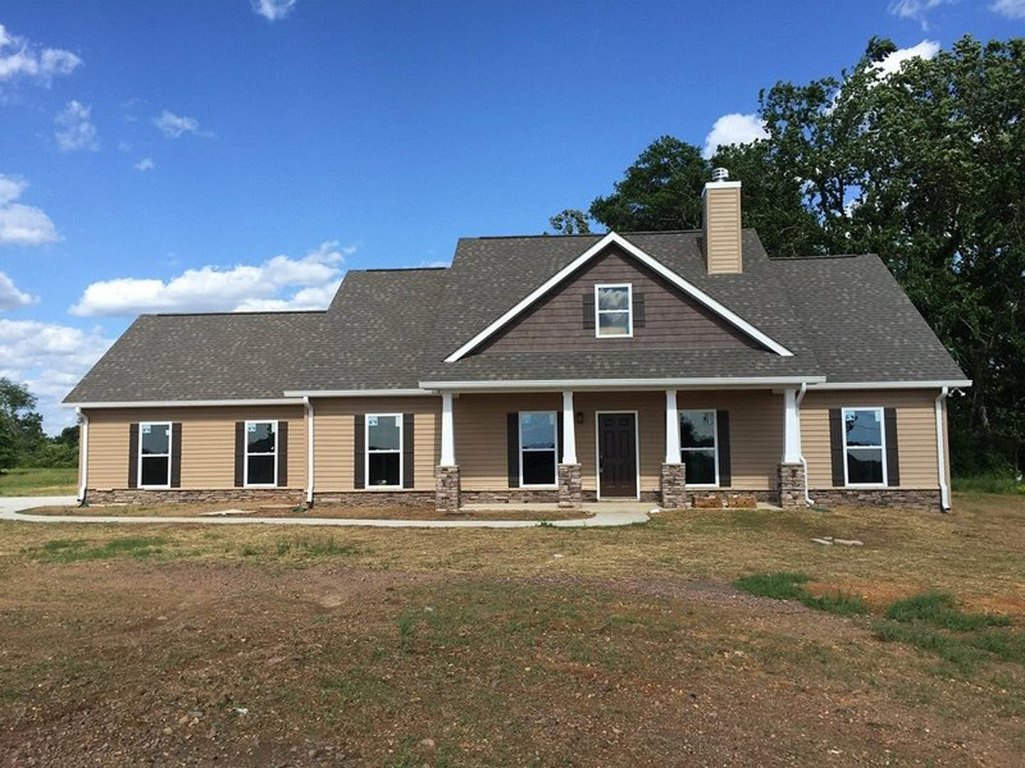 Brown-roofed house with white-framed windows and black front door, surrounded by a large grassy yard, trees, and cloudy sky; one window shows broken glass.