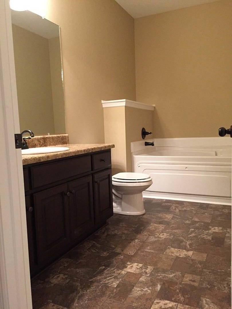 Brown tile bathroom with white bathtub, white toilet, wood vanity cabinet, and chrome faucet