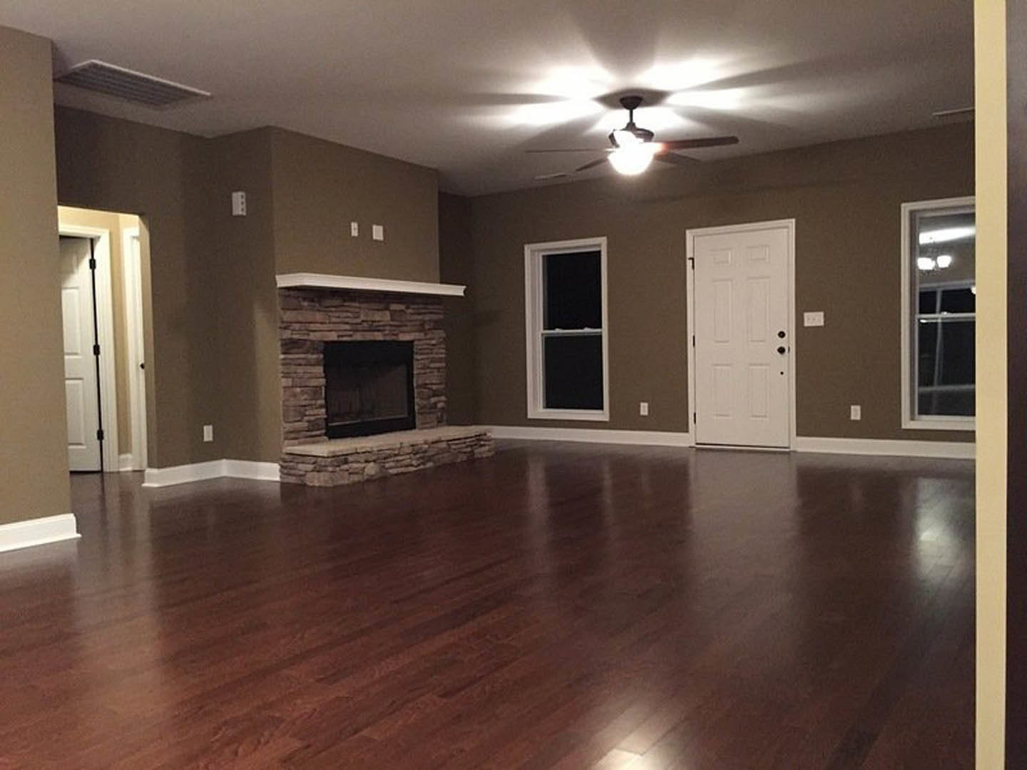 Living room with hardwood floors, stone fireplace centered on wall, white-framed window, white door with black knobs, ceiling fan, neutral walls