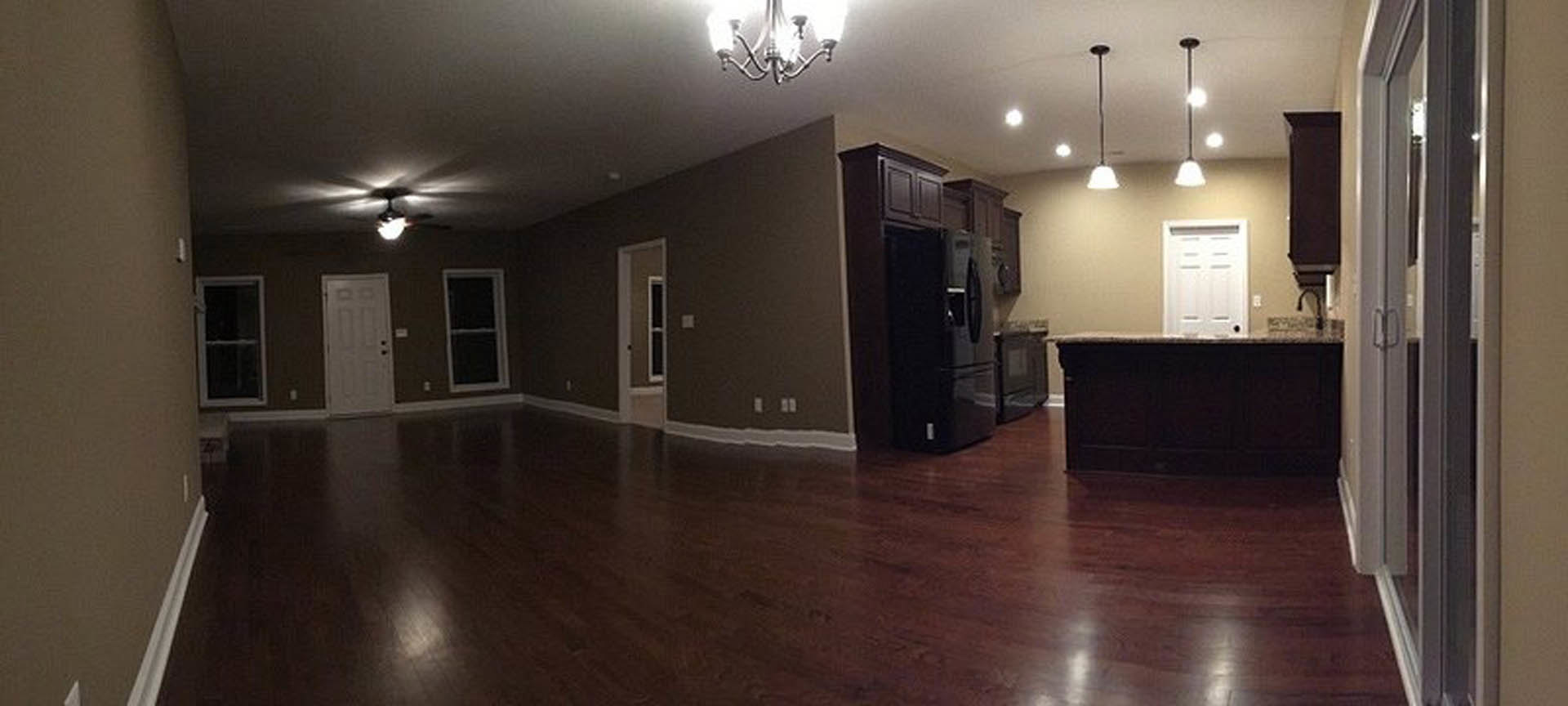 Spacious living room with dark hardwood floors, white walls, recessed ceiling lights, and a black refrigerator visible near the kitchen area