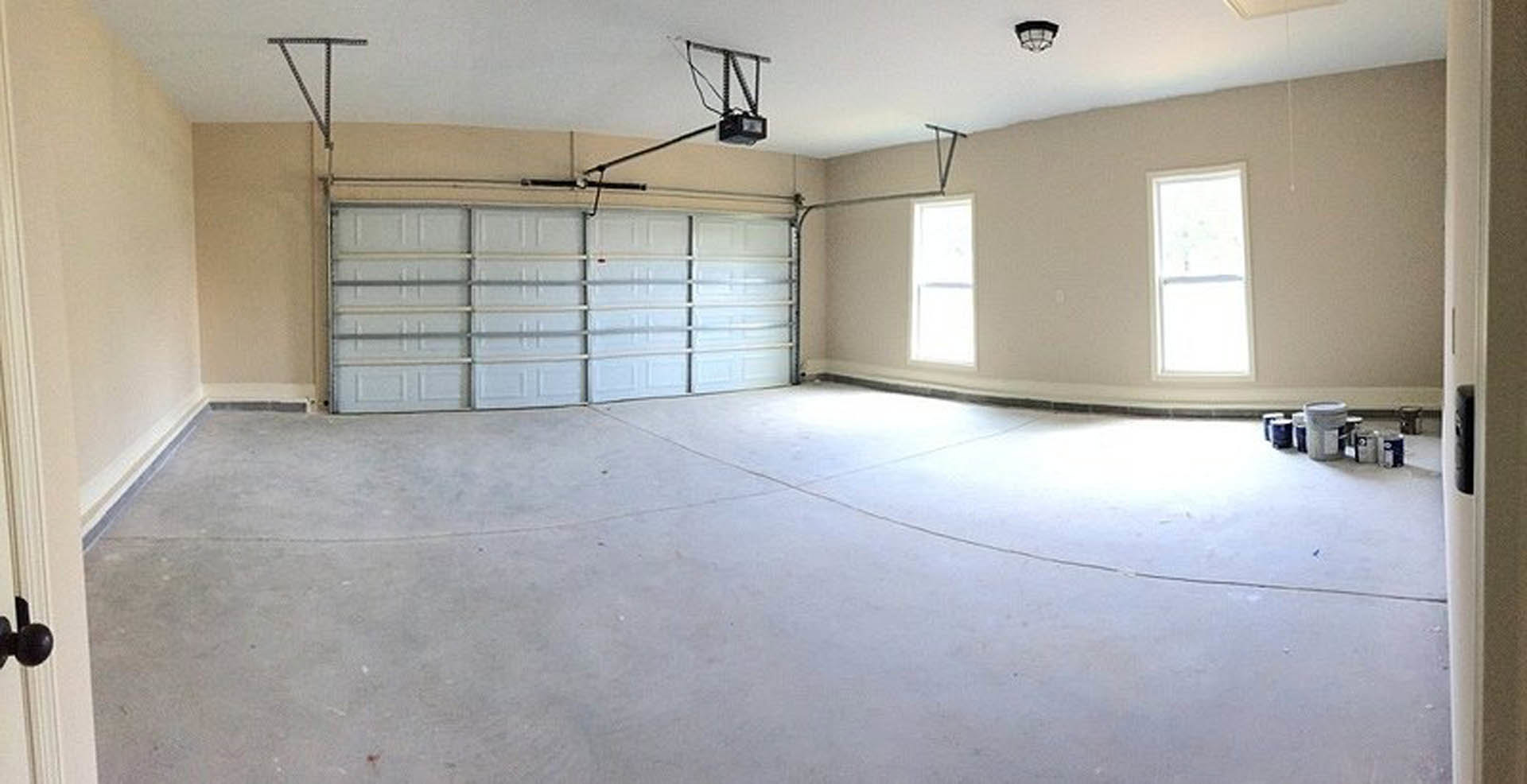 Spacious garage with white plaster walls, tile flooring, ceiling-mounted black and white light fixture, window letting in natural light, built-in shelves beside garage door