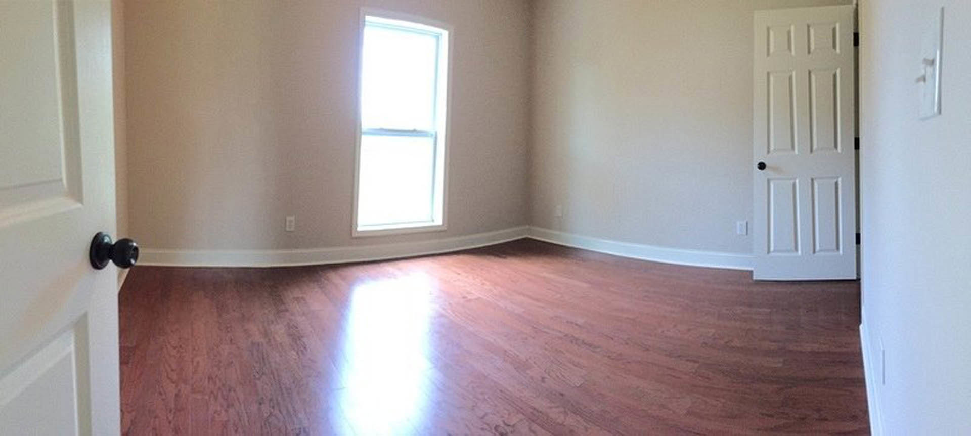 Sunlit hardwood floor in a room with white walls, large window, and white door featuring a black knob