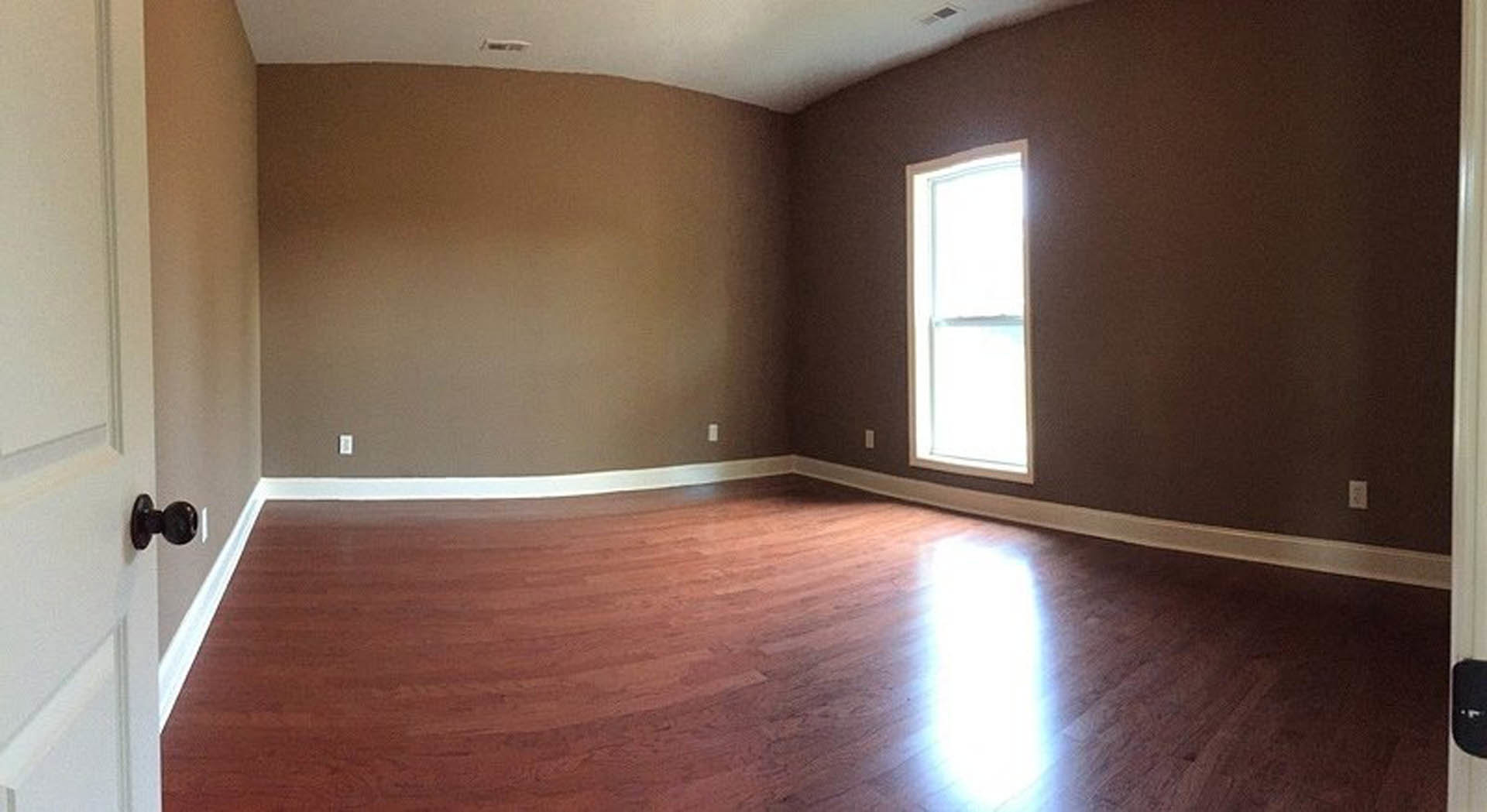 Sunlit room featuring a large window, brown plaster walls, and polished hardwood flooring with light reflecting across the surface.