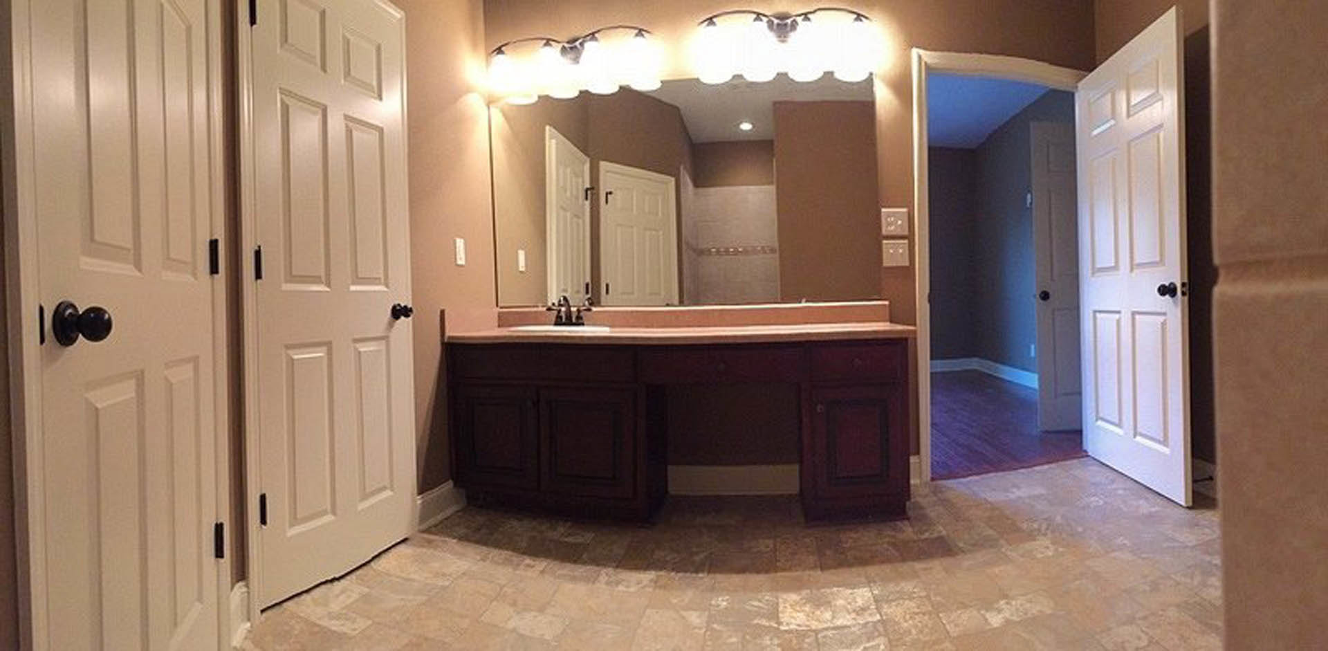 Bathroom with expansive wall mirror, brown tile floor bordered in white, white cabinetry, modern sink and tap, ceiling lights, white door visible in foreground