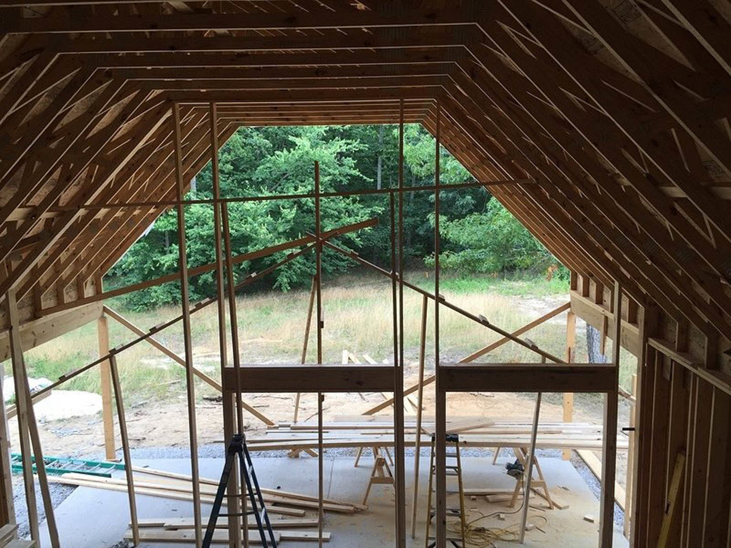 Exposed wood framing and beams in a partially constructed room, ladder leaning against a post, sunlight filtering through open walls, trees visible outside