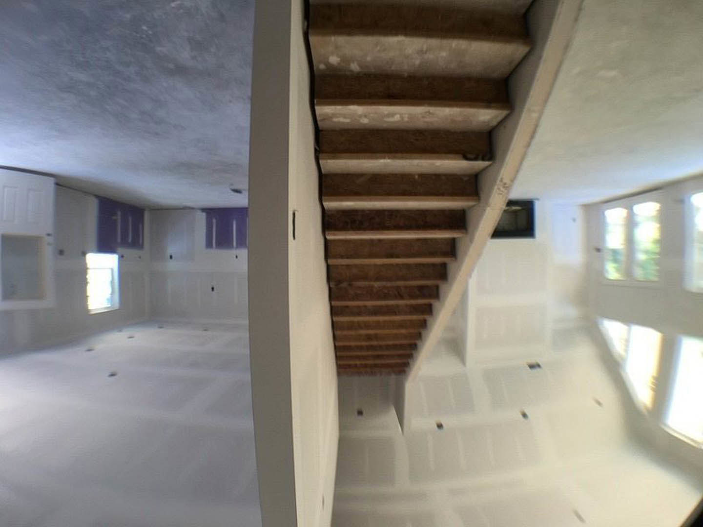 Wood staircase with white risers and handrail in a bright room, sunlight streaming through a large window onto plaster walls and ceiling beams, door visible in background.