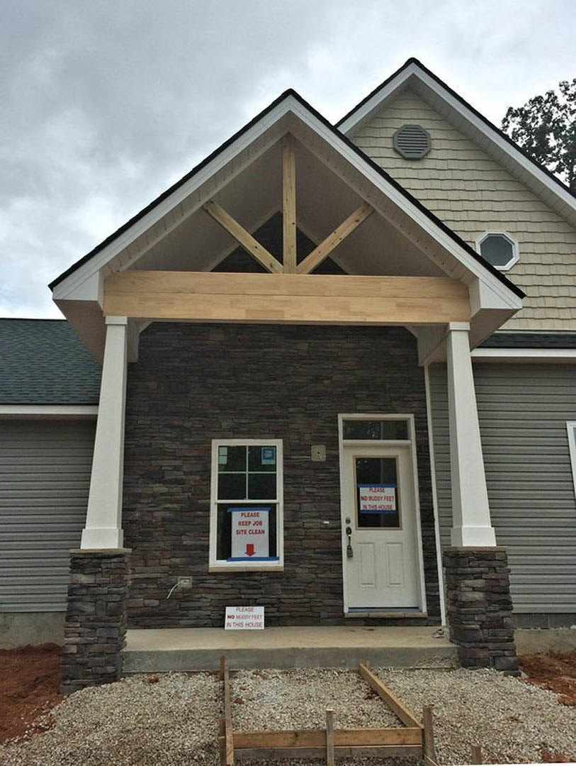 Modern home exterior with exposed wood beam above covered porch, white door displaying a sign, grey wall vent, large window, and construction materials including gravel and lumber