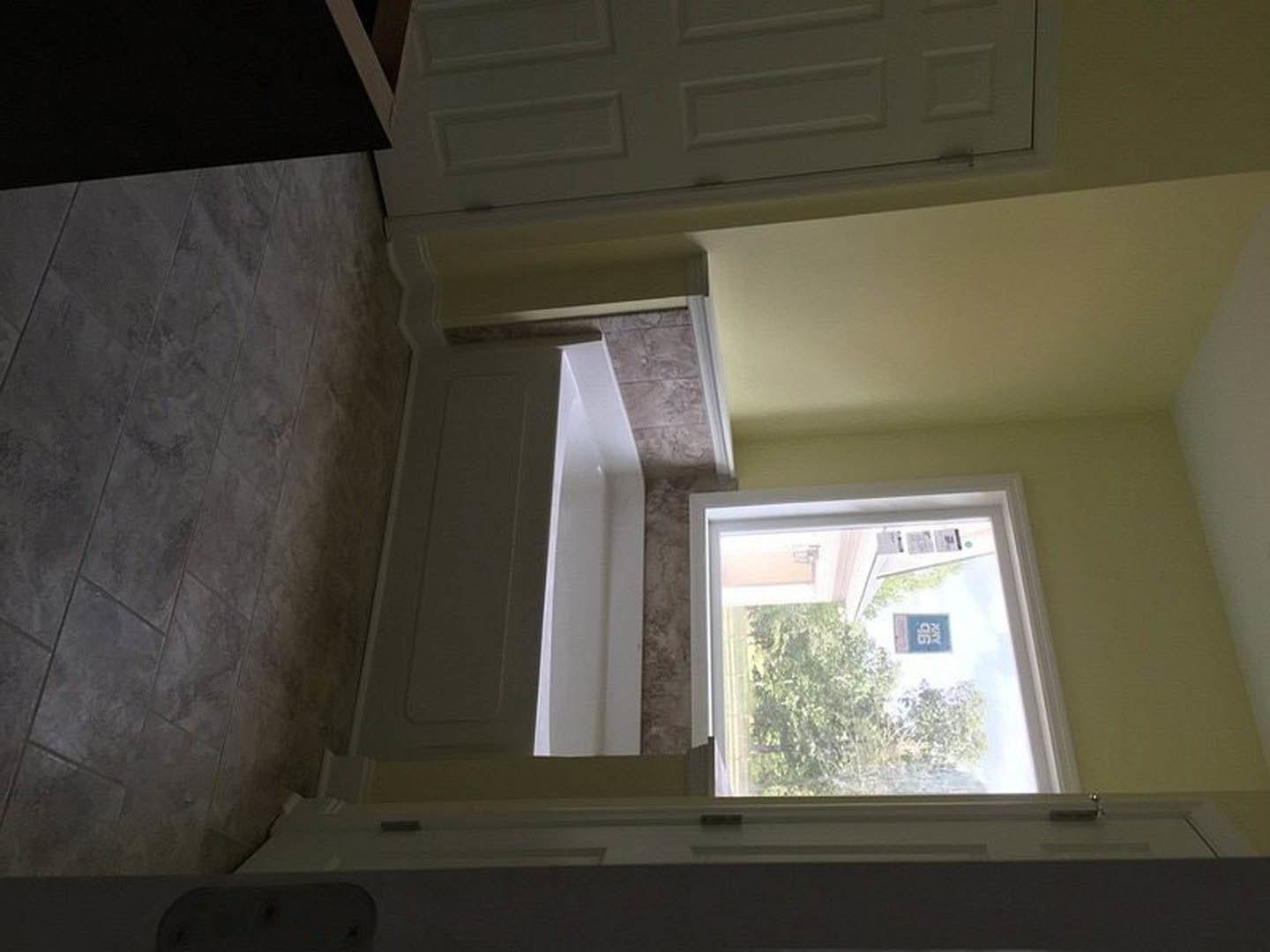 White bathtub beneath a large window, sunlight streaming onto pale plaster walls, blue and white sign visible outside, white door partially in frame