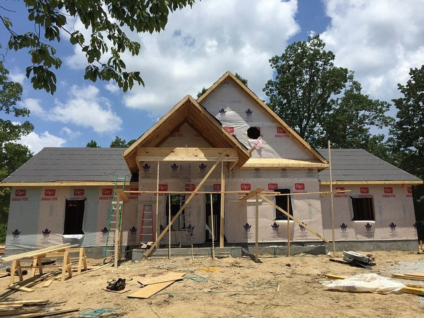 Two-story wood-framed house under construction with metal scaffolding, ladder leaning against exterior, exposed plywood sheathing, surrounded by dirt and scattered lumber, cloudy