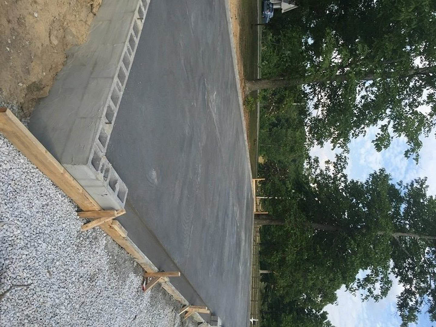 Concrete foundation slab bordered by cement blocks, adjacent to a paved road lined with leafy trees, wooden framing materials nearby, and a concrete curb featuring square holes