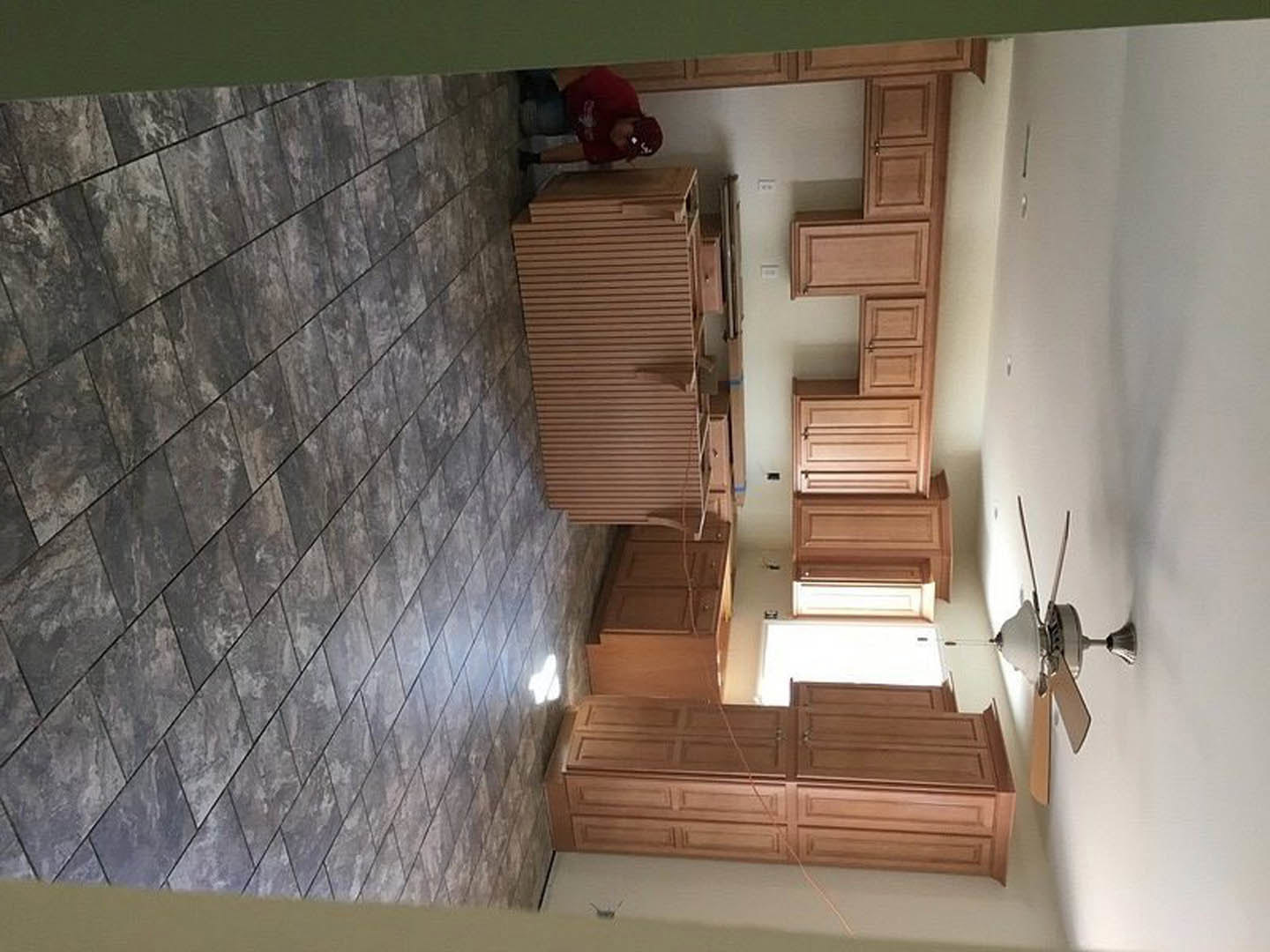 Person in red shirt standing in modern kitchen with wooden cabinetry, brown accent wall featuring black lines, grey tile flooring illuminated by overhead lighting, and white wall