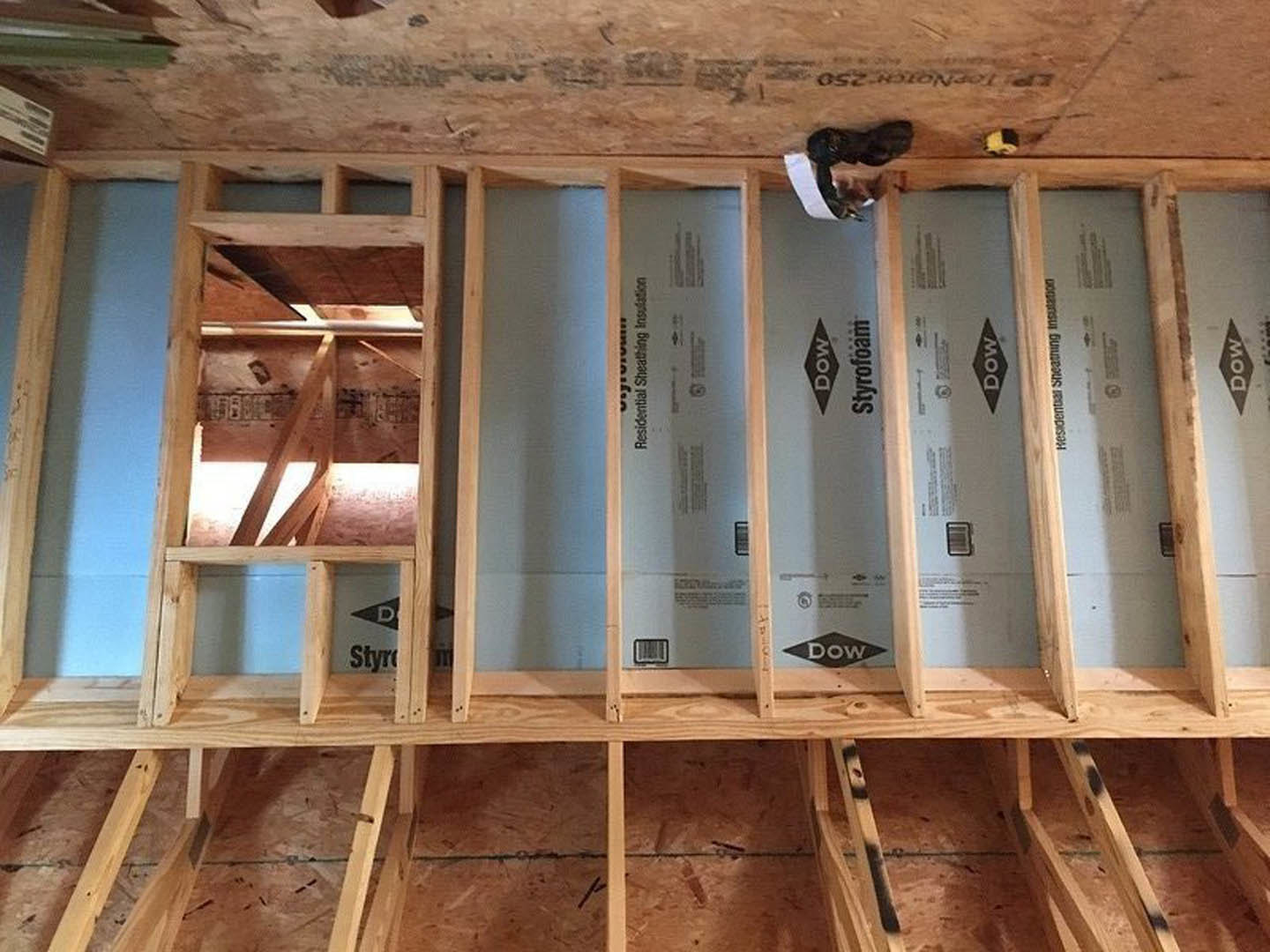 Exposed wooden framing and beams in a partially constructed room, person in a cap standing on subfloor, black dog nearby, unfinished walls and insulation visible