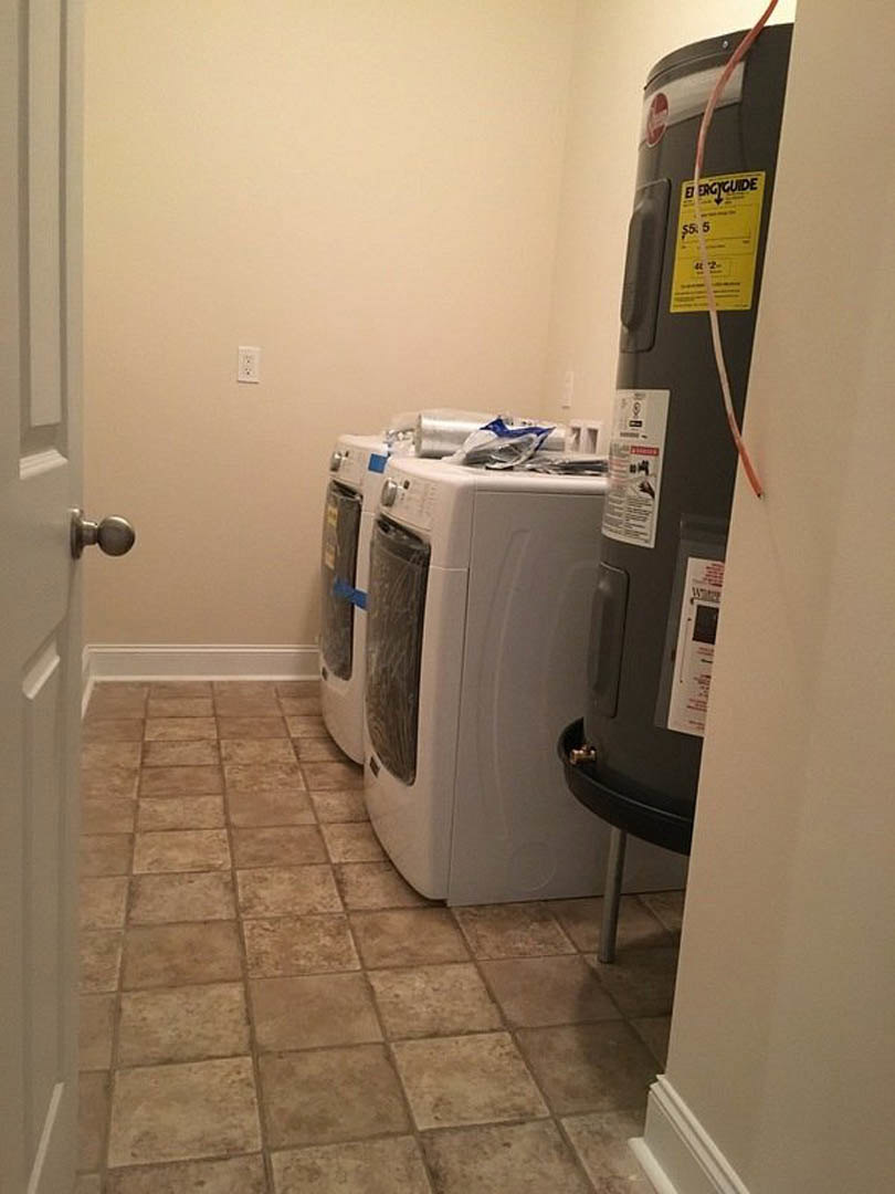 White washing machine beside cylindrical water heater on light tile floor, adjacent to beige wall and wooden door, yellow caution sign visible.