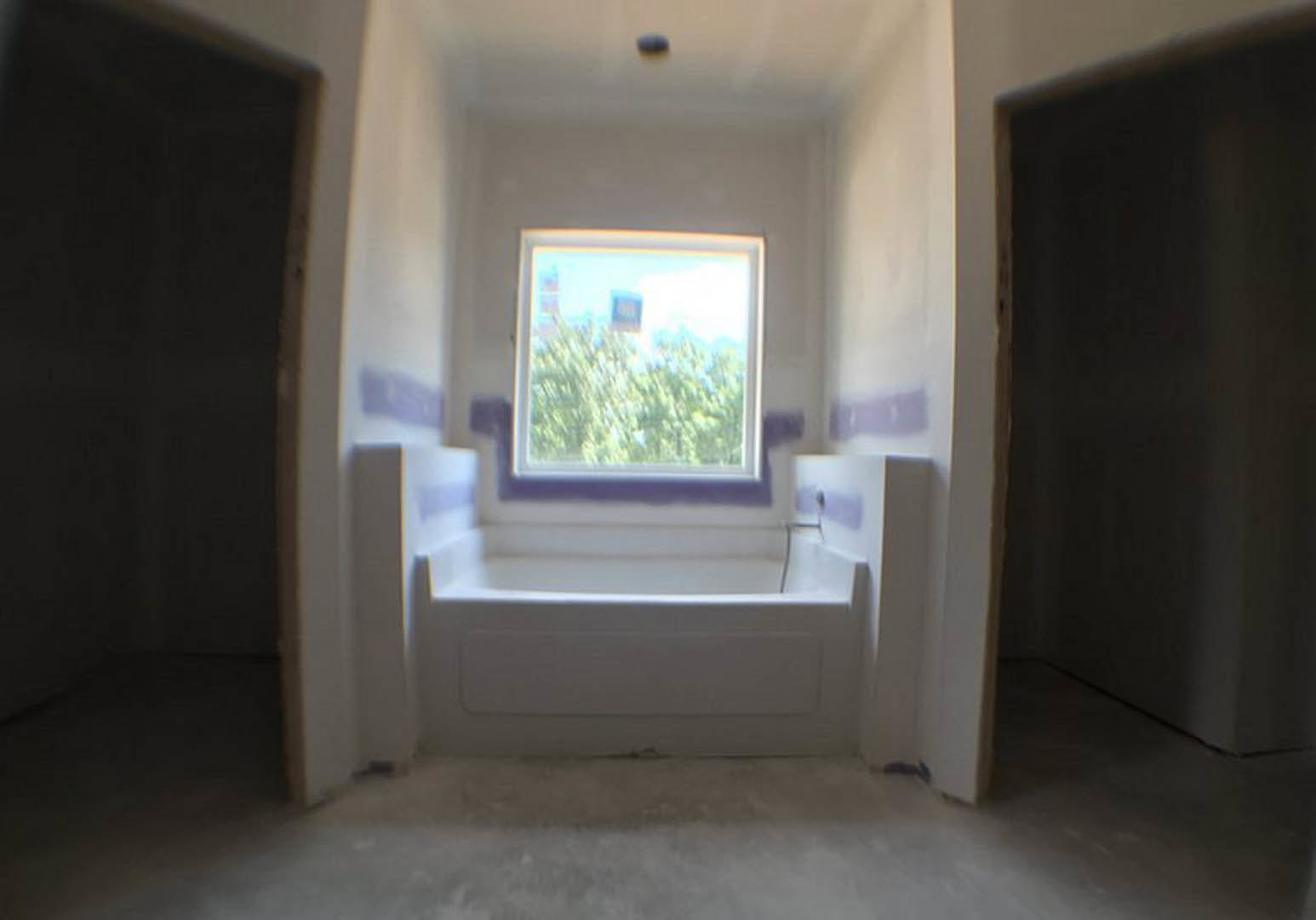 White freestanding bathtub with purple frame set near a window, blurred trees visible outside, plaster walls, light flooring, partial view of a hand in foreground.