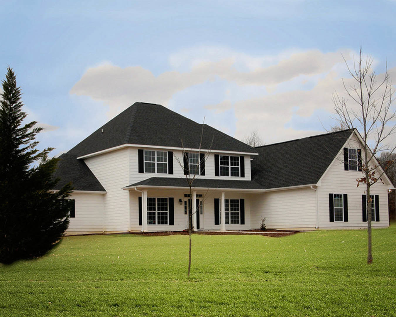 White two-story house with black roof, expansive green lawn, leafless tree in foreground, large windows, partly cloudy sky overhead