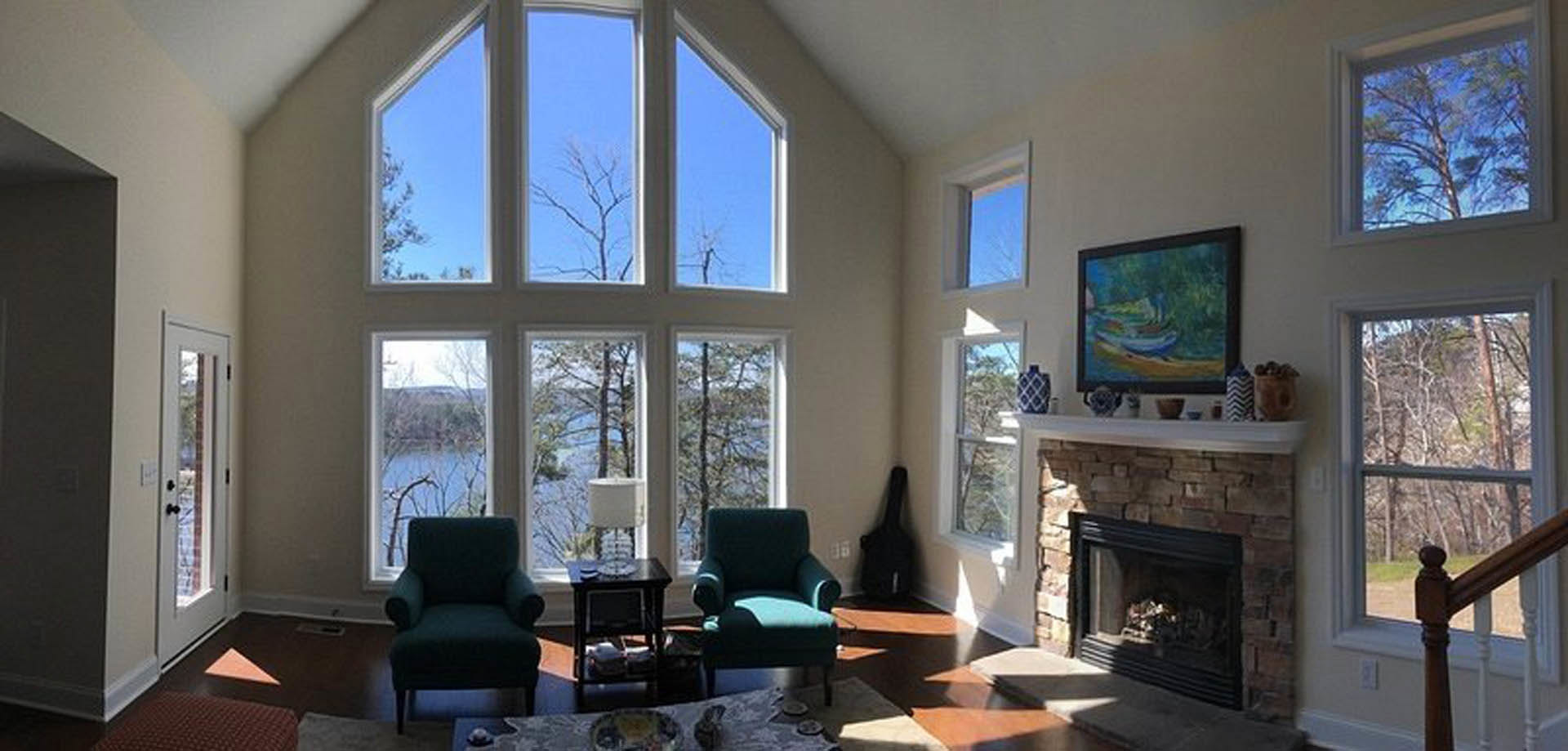 Living room with stone fireplace, green armchair near window, blue chair with matching cushion, abstract painting on wall, wooden railing in foreground