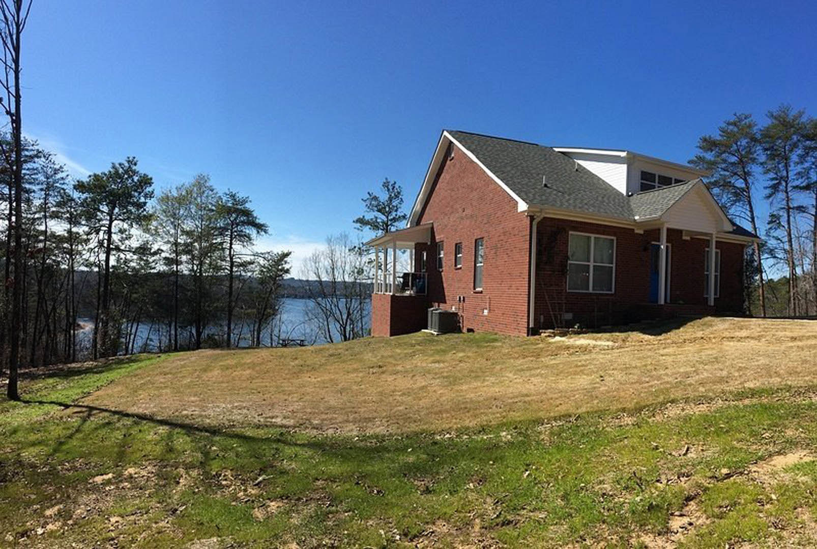 Brick house with blue door and white-framed windows, situated on a grassy hill overlooking a lake, surrounded by trees under a clear sky