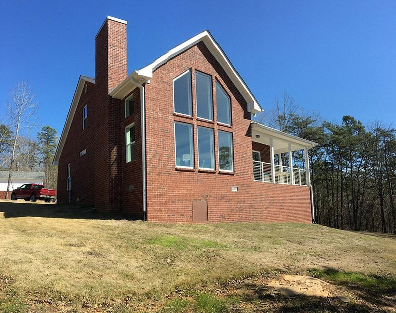 Brick house with covered porch, white door, blue-trimmed window, red truck parked on grassy area behind, trees in background