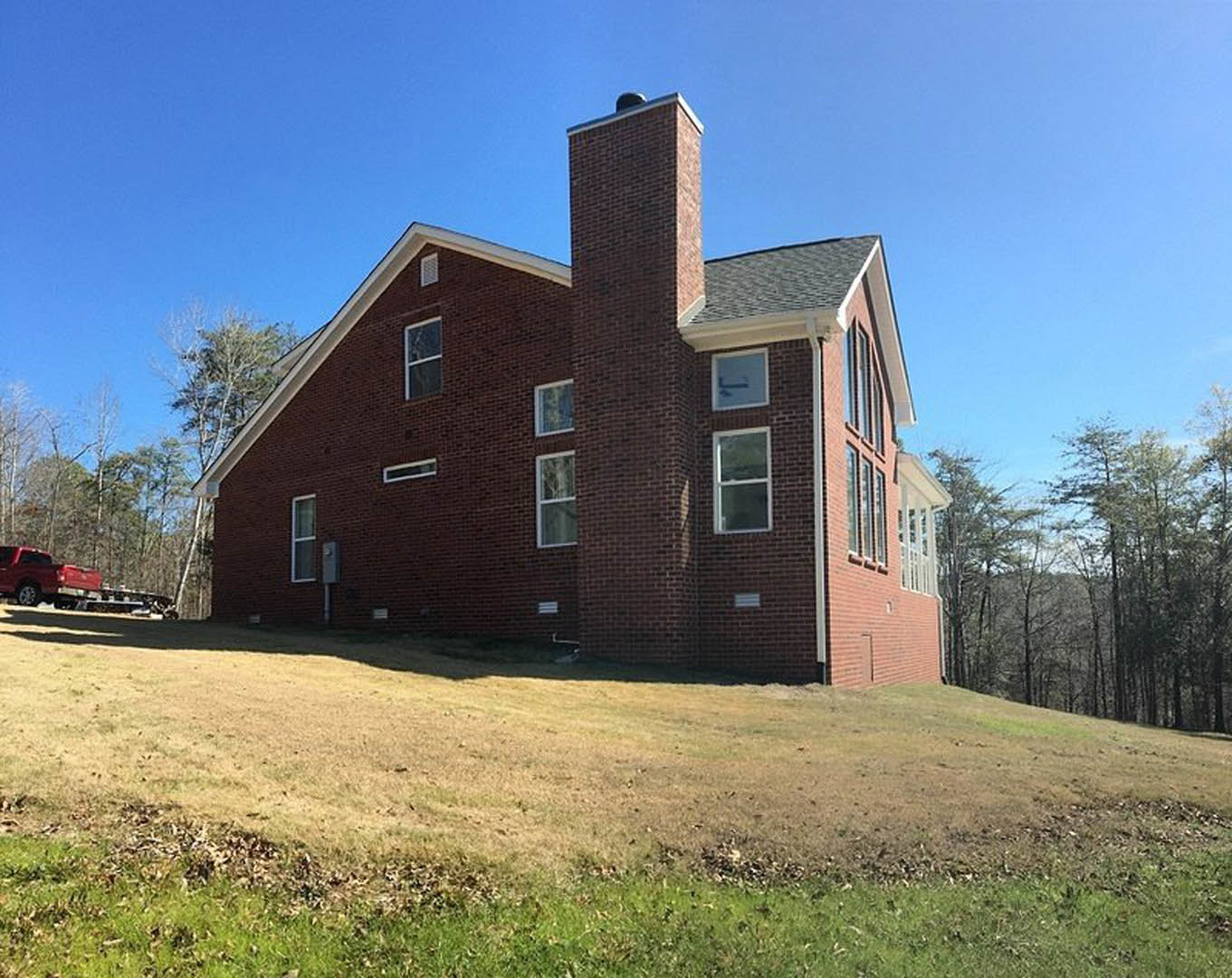 Brick home with chimney situated on grassy hill, surrounded by mature trees, large windows visible on facade, red truck parked on nearby road under blue sky