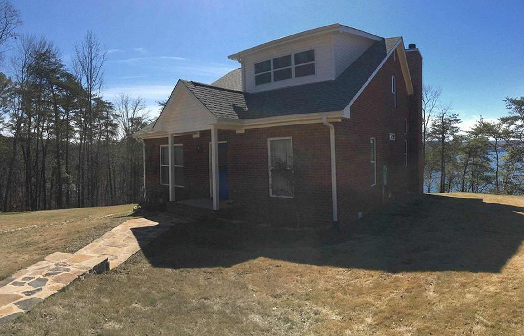 Red brick house with dormer windows, stone walkway leading through grassy lawn, lake visible behind the home, leafy plant in front window