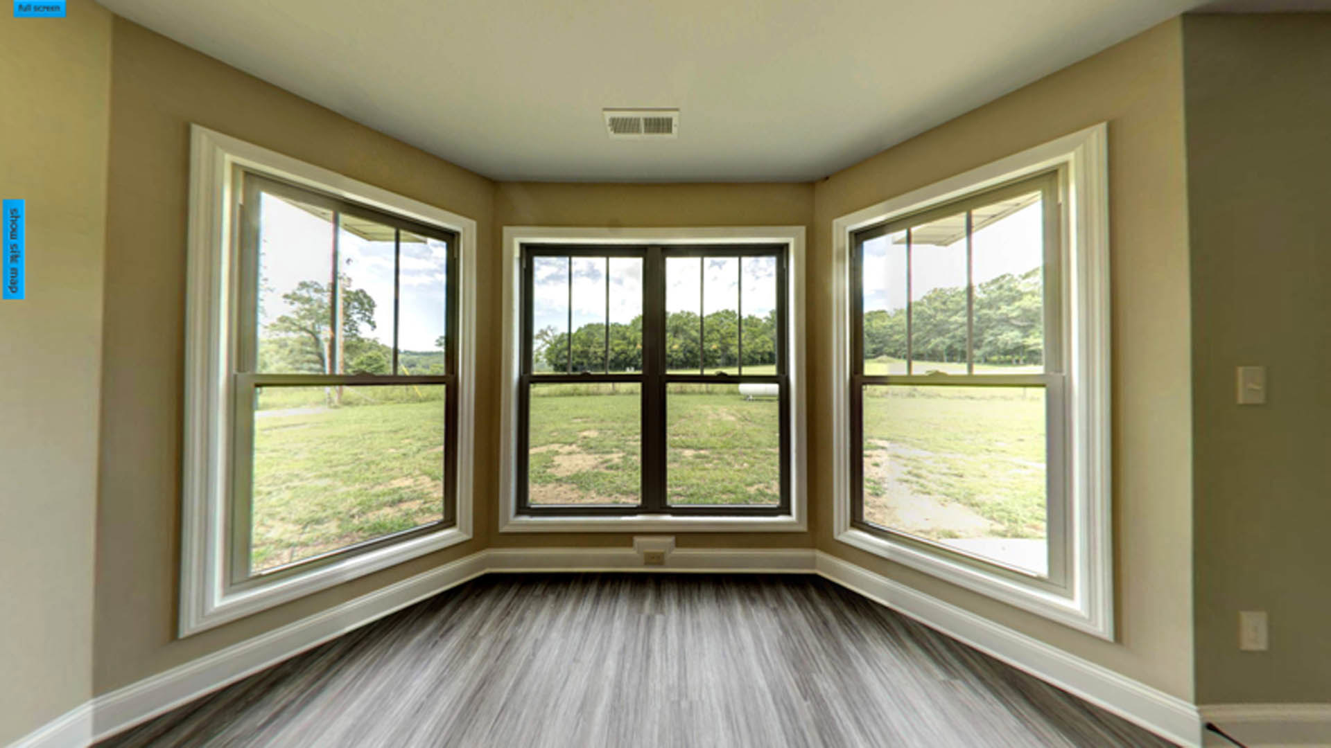 Sunlit room with large windows, wood flooring, white walls, light switch, wall vent, and grassy field visible outside