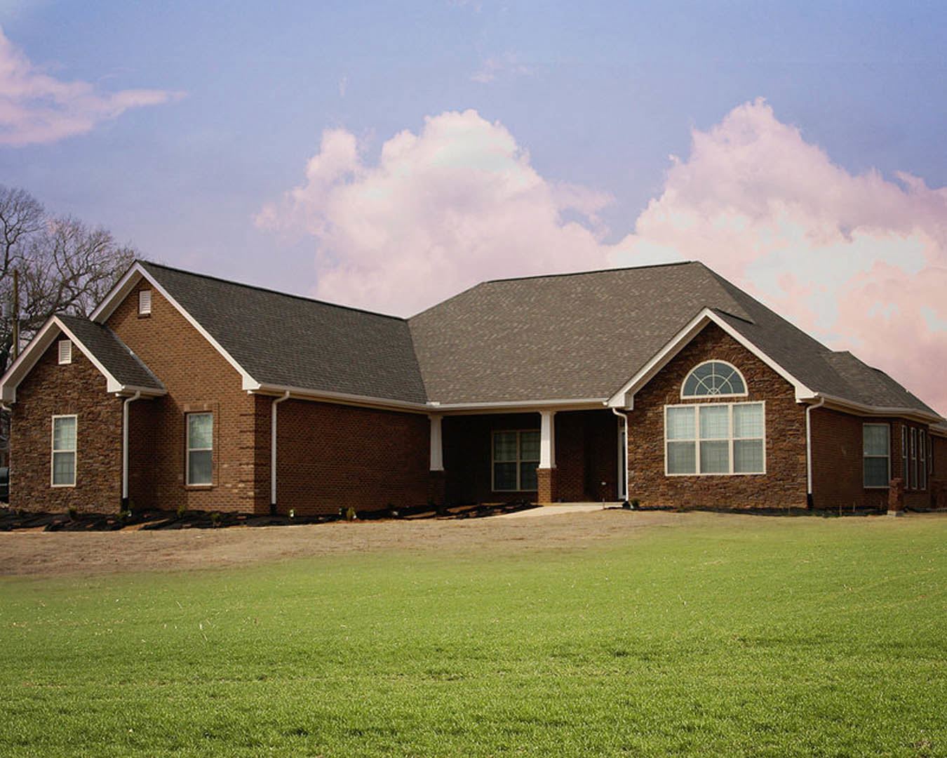 Brick exterior home with white columns, large windows, manicured green lawn, and a shingled roof framed by mature trees under a partly cloudy sky