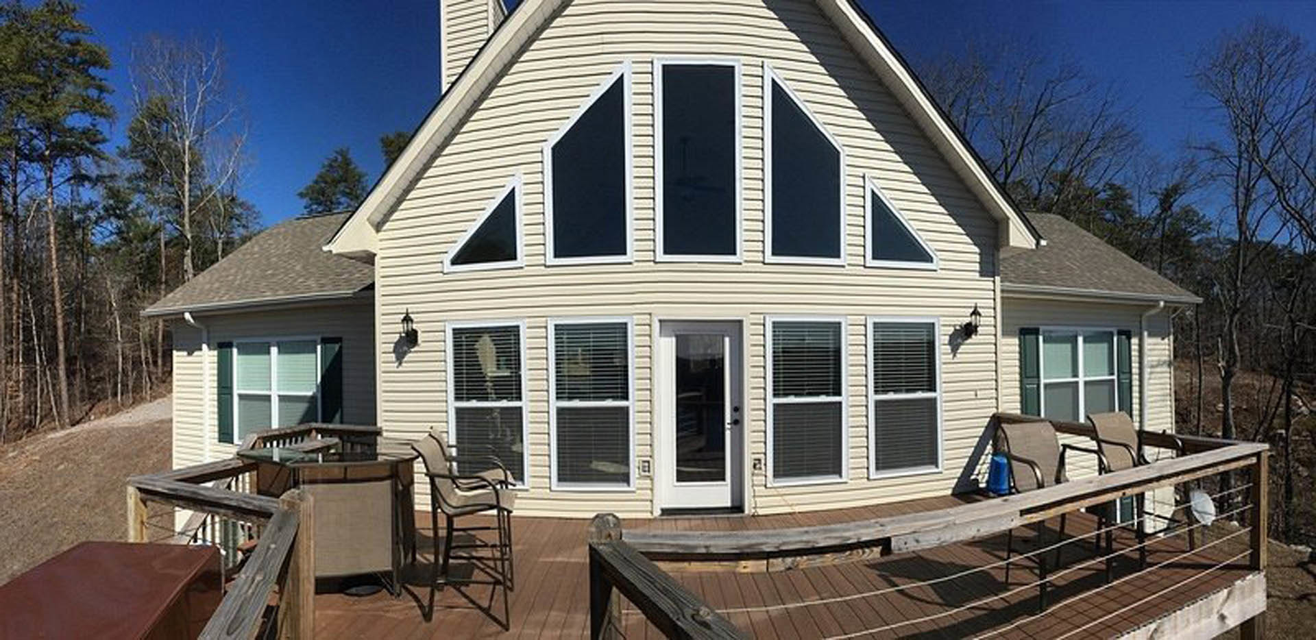 Modern home with light siding, white framed windows, and a white door, featuring a wooden deck with a bench and chair, stairs leading to a stone patio, surrounded by trees and blue